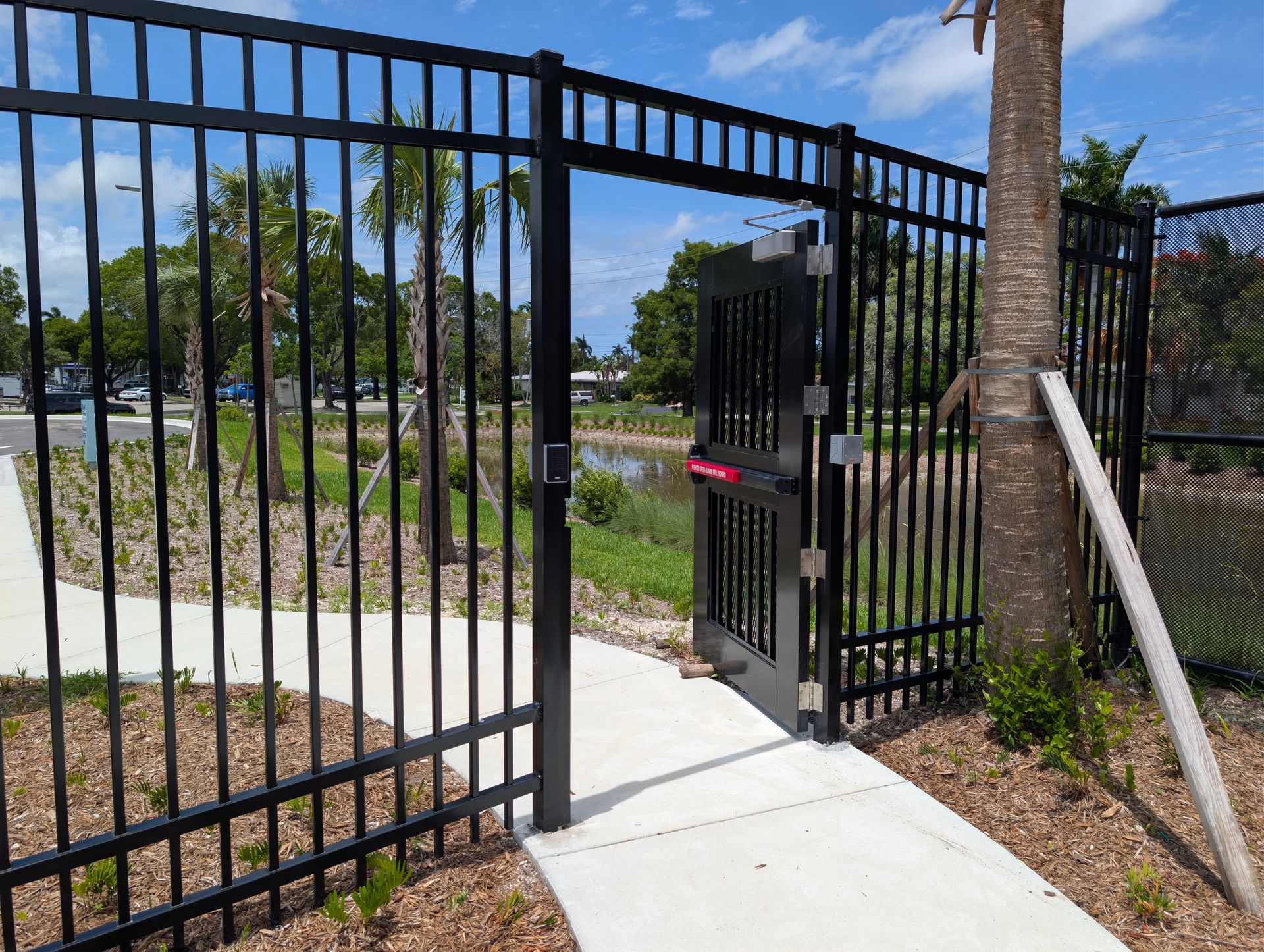 Black metal gate opening onto a sunny walkway in a landscaped park area.