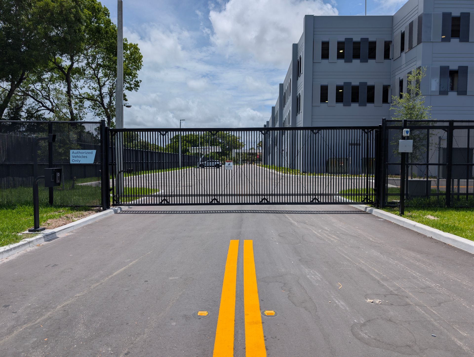 Closed black gate blocking a road leading to a modern building under a partly cloudy sky
