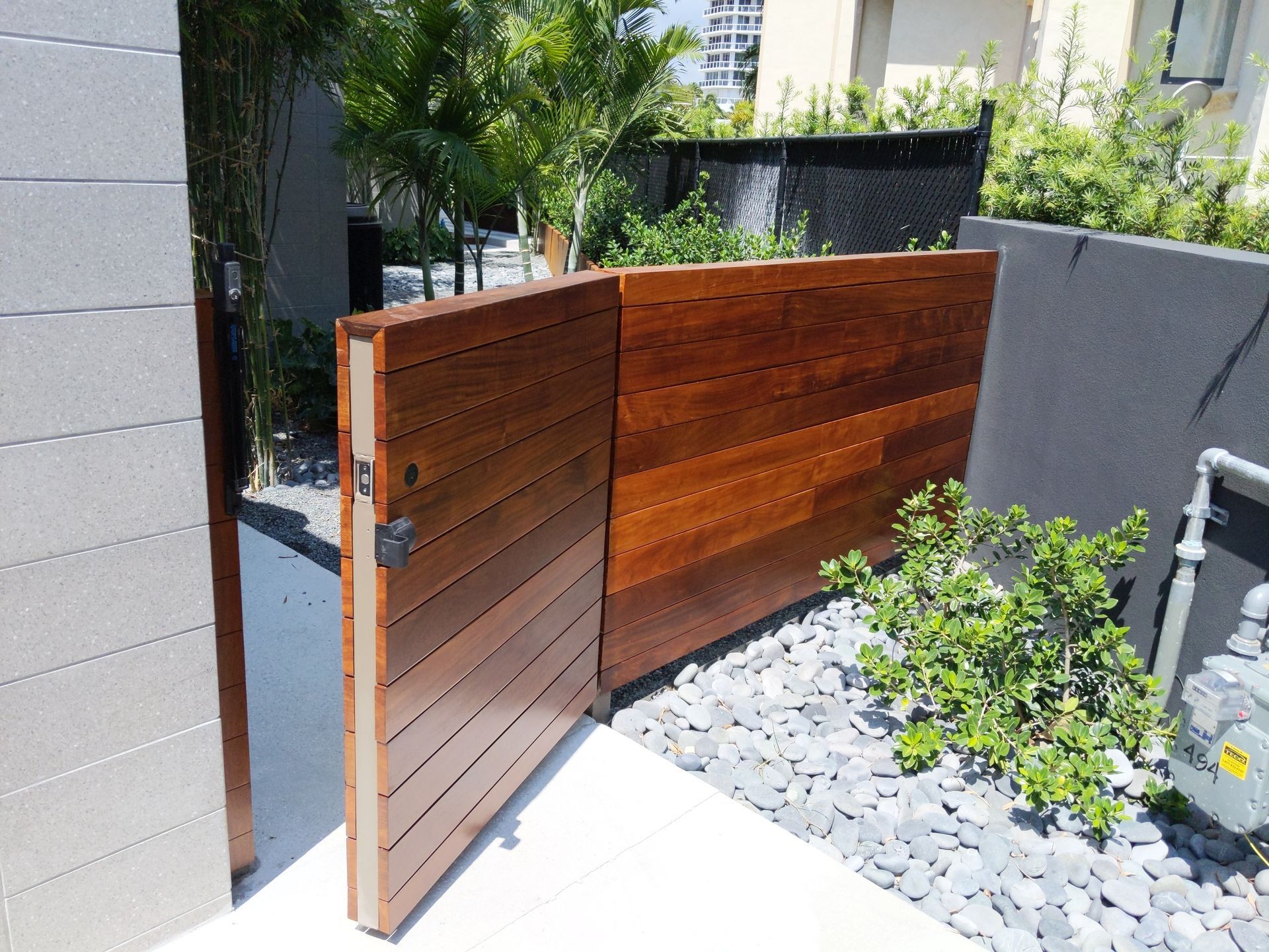 Wooden garden gate beside a gray wall, with gravel landscaping and greenery in bright sunlight