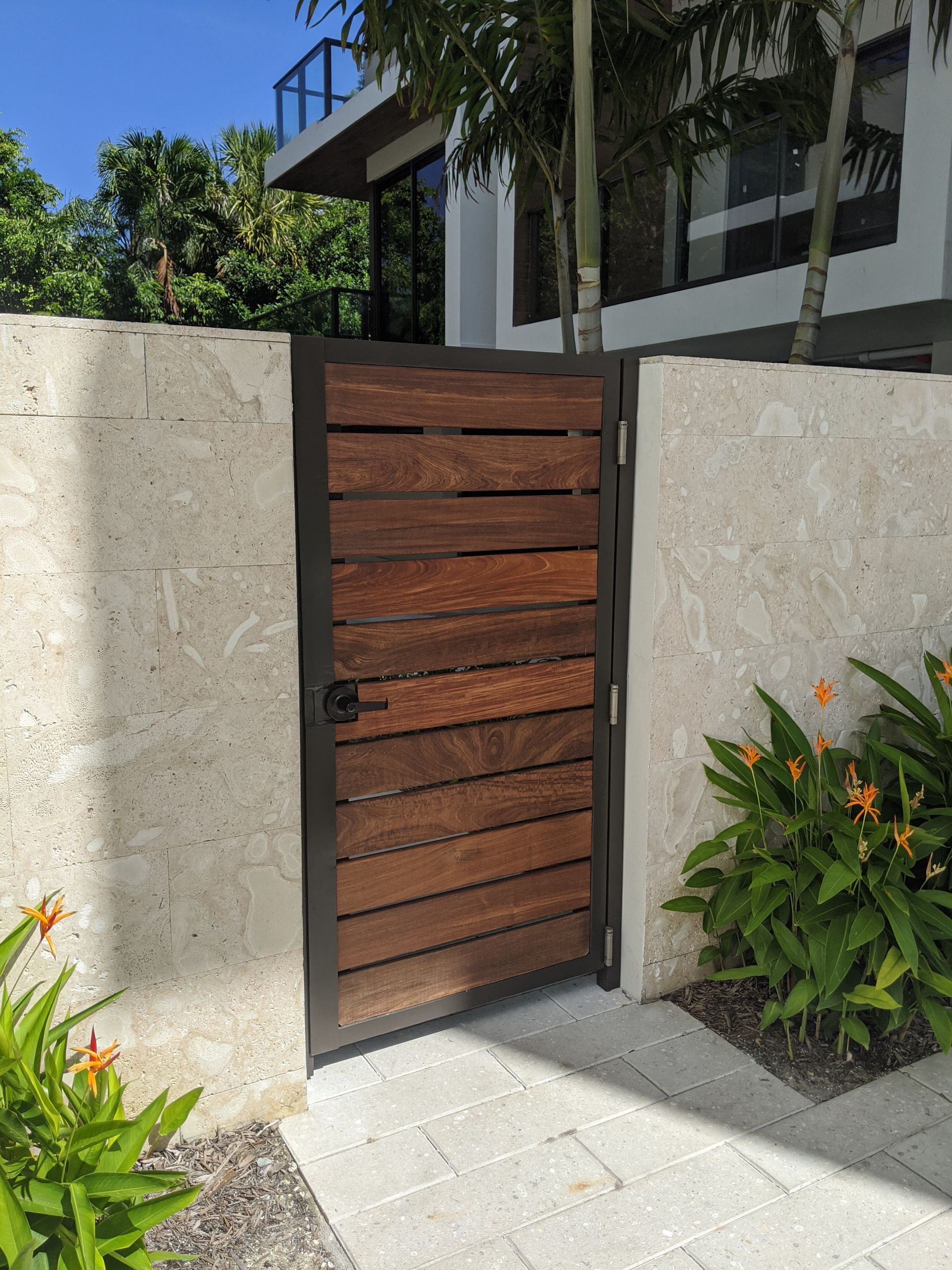 Wooden pedestrian gate between stone walls beside tropical plants and a modern house entrance.