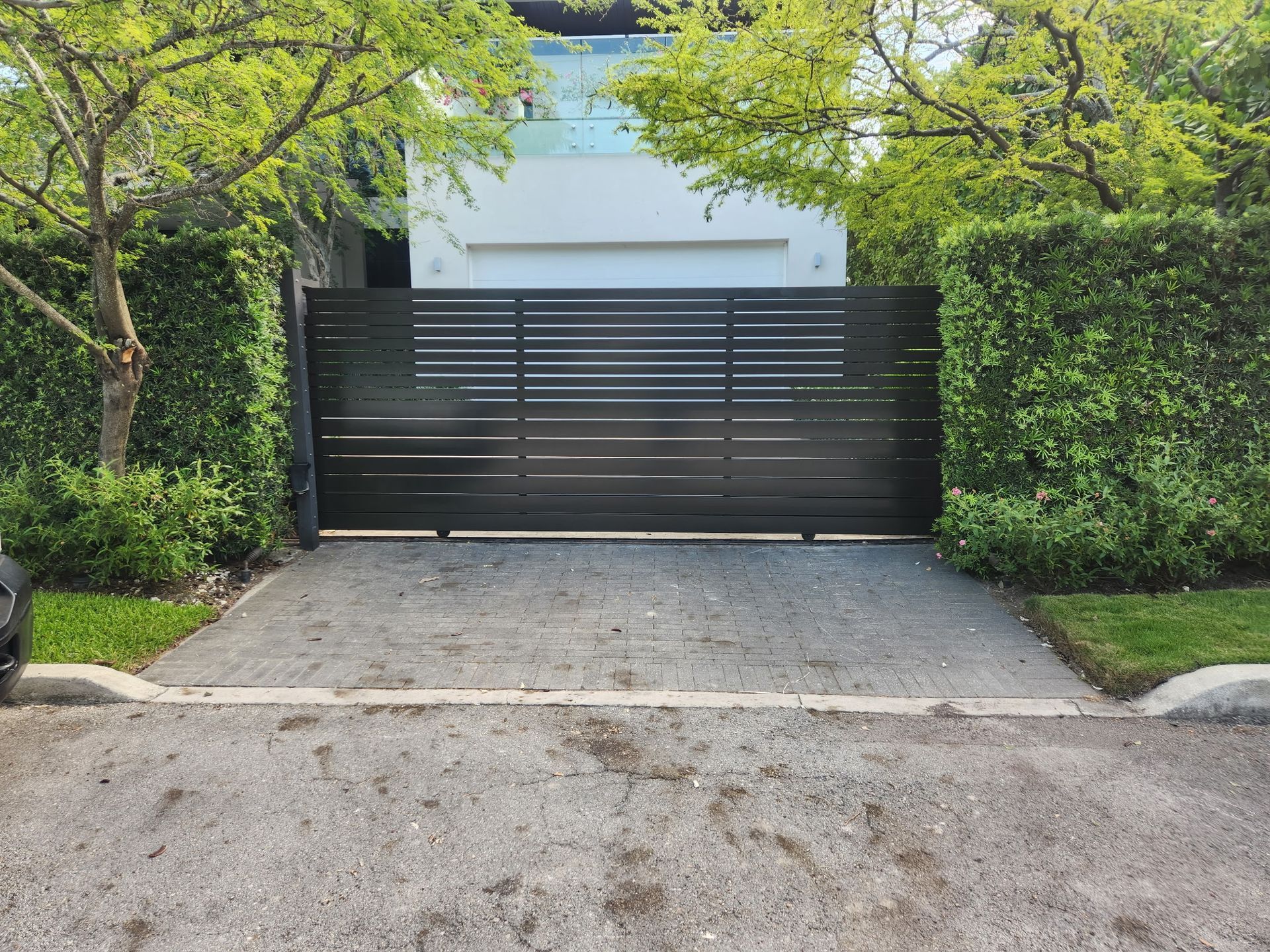 Black horizontal-slat gate at the end of a driveway, framed by green hedges and trees