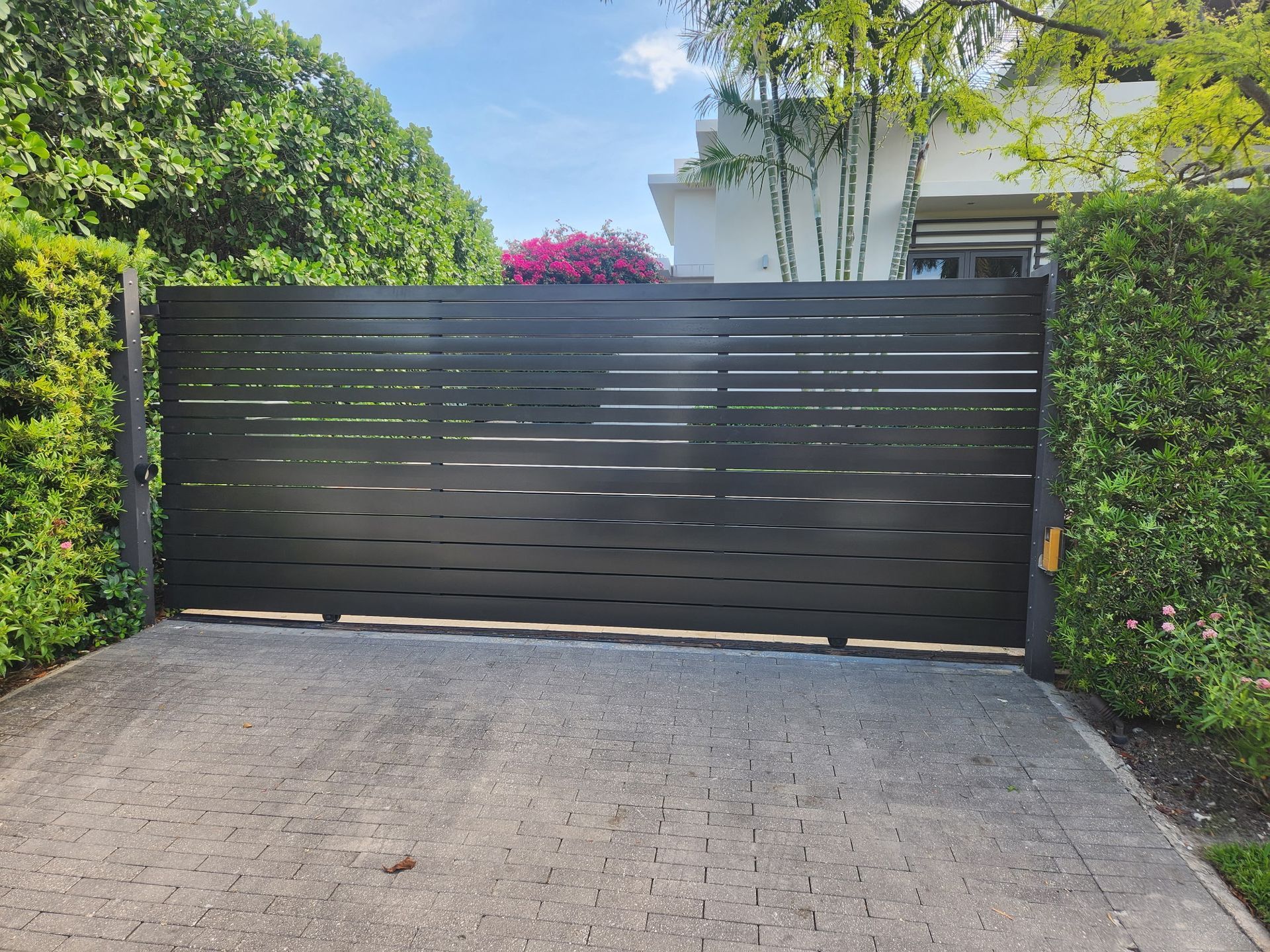 Black sliding driveway gate in front of a hedged home, with a paved driveway and trees behind it.