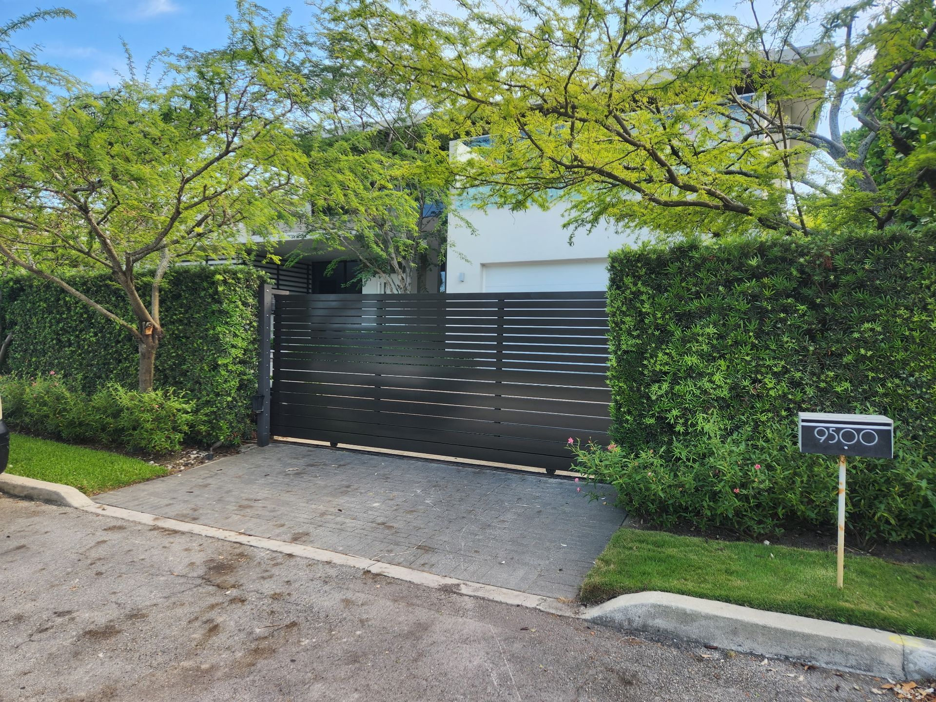 Modern house entrance with black gate, hedge, and driveway under green trees