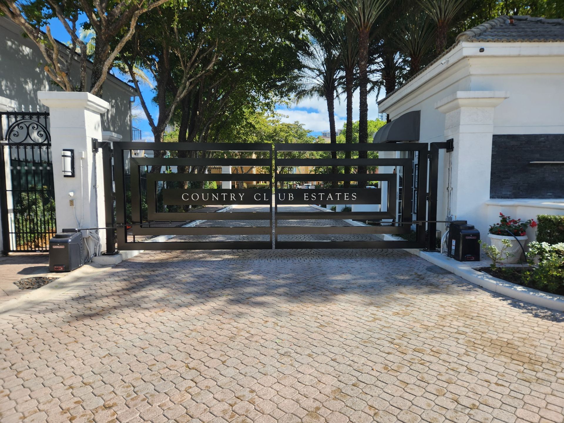 Black gate entrance with stone pillars and palm trees, leading to a cobblestone driveway