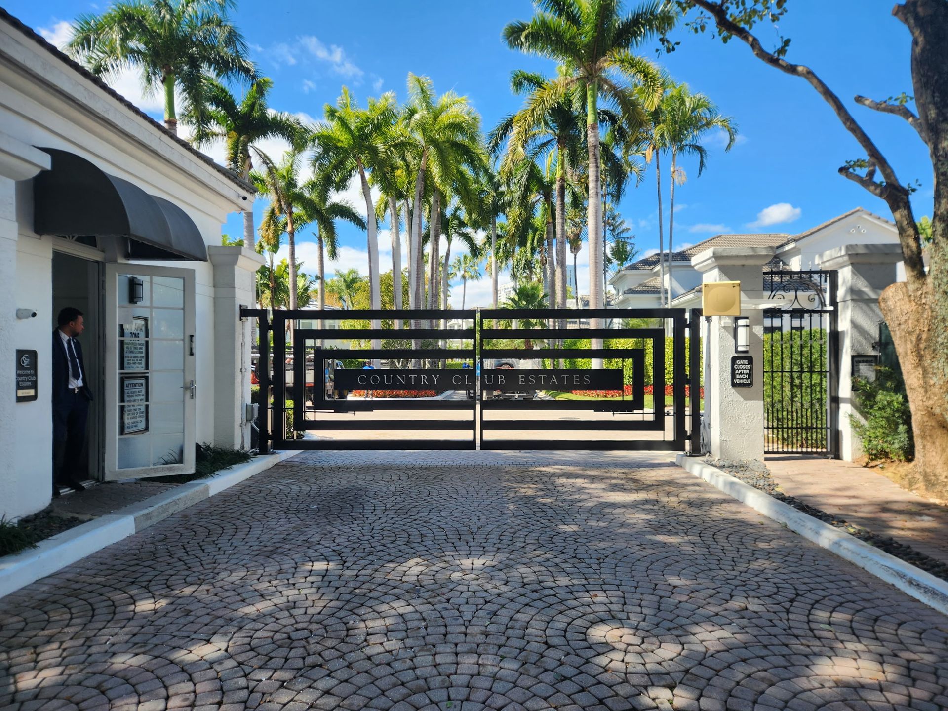 Gated driveway entrance with black gate, palm trees, and white buildings under a blue sky