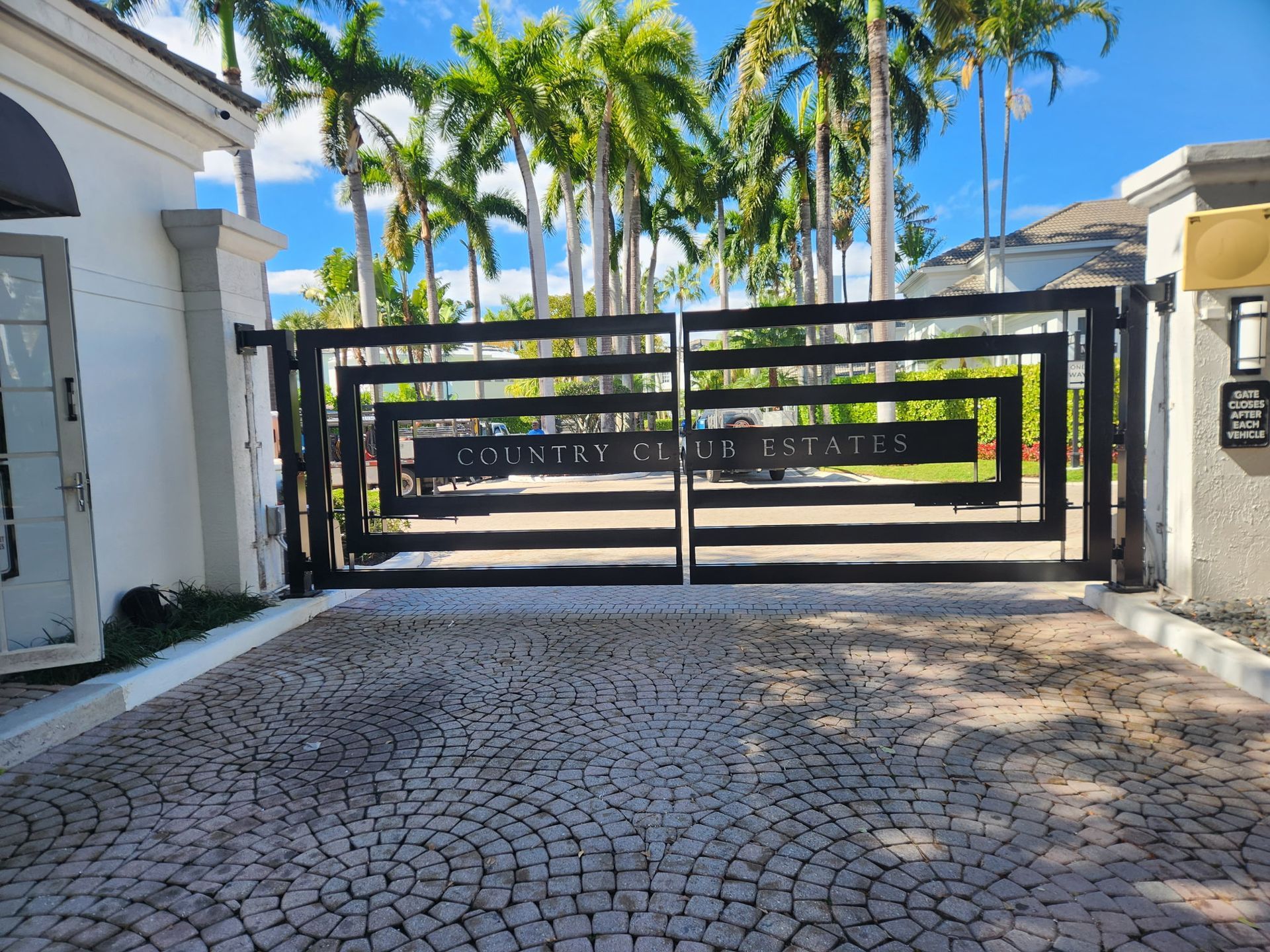 Closed black gate at a palm-lined entrance with cobblestone driveway and stone pillars