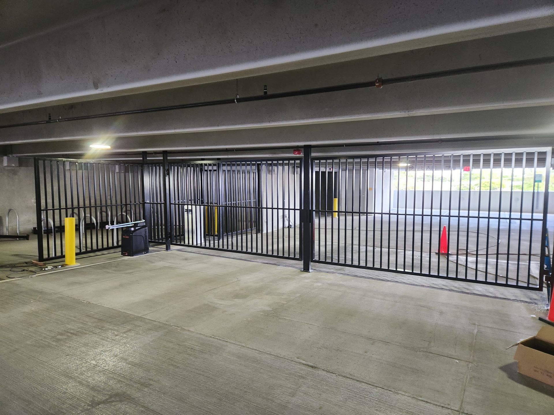 Closed black metal gate in a parking garage, with yellow bollards and orange cones nearby