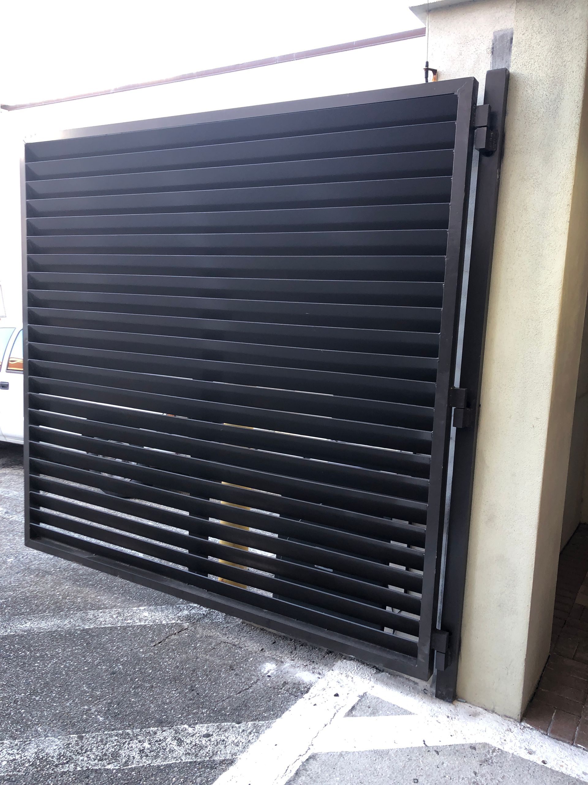 Black slatted metal driveway gate partially open beside a concrete wall