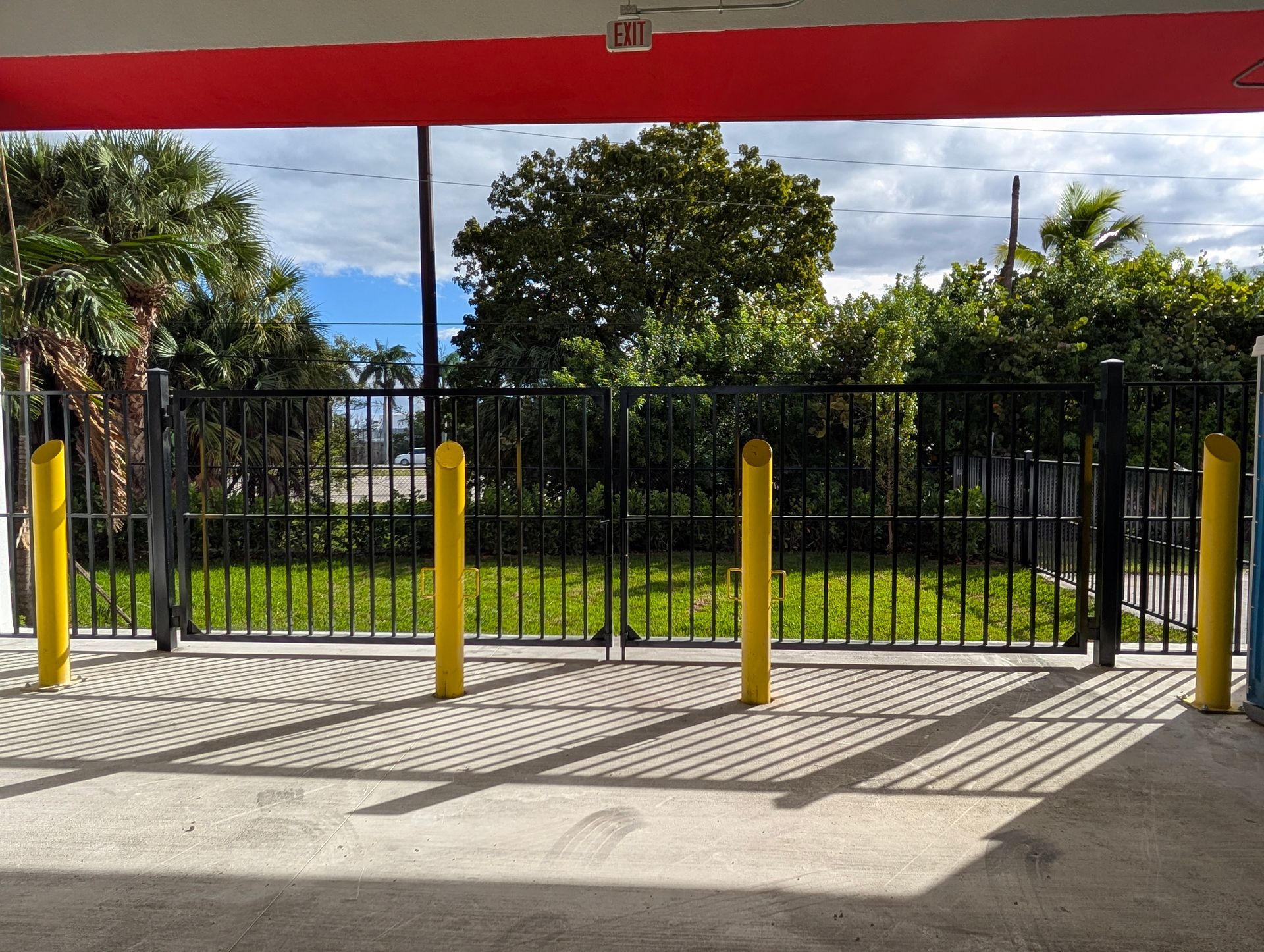 Black metal fence with yellow bollards beside a sunlit driveway and grassy yard under a red awning
