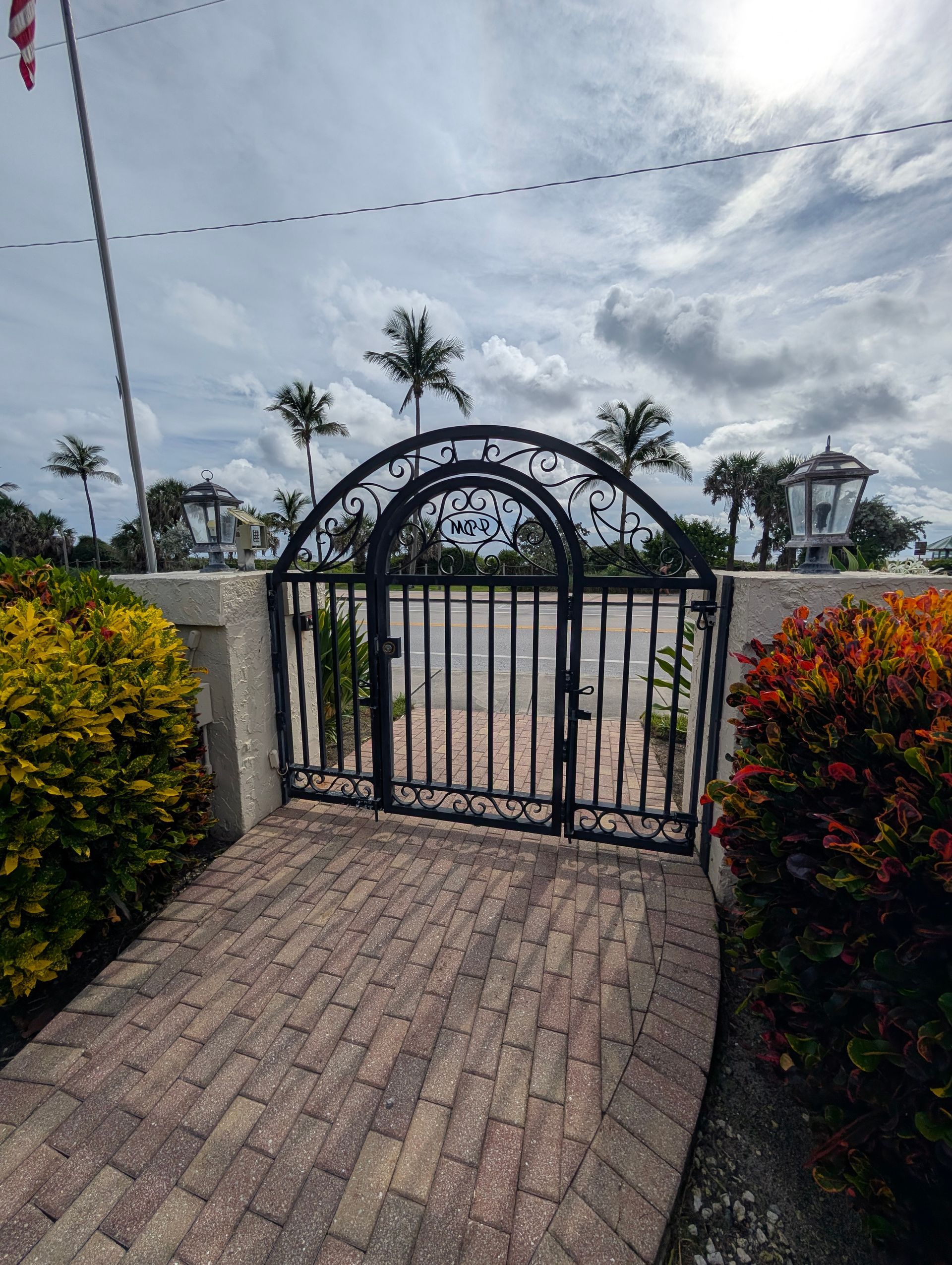 Black iron gate at a brick walkway entrance with palm trees and cloudy sky beyond