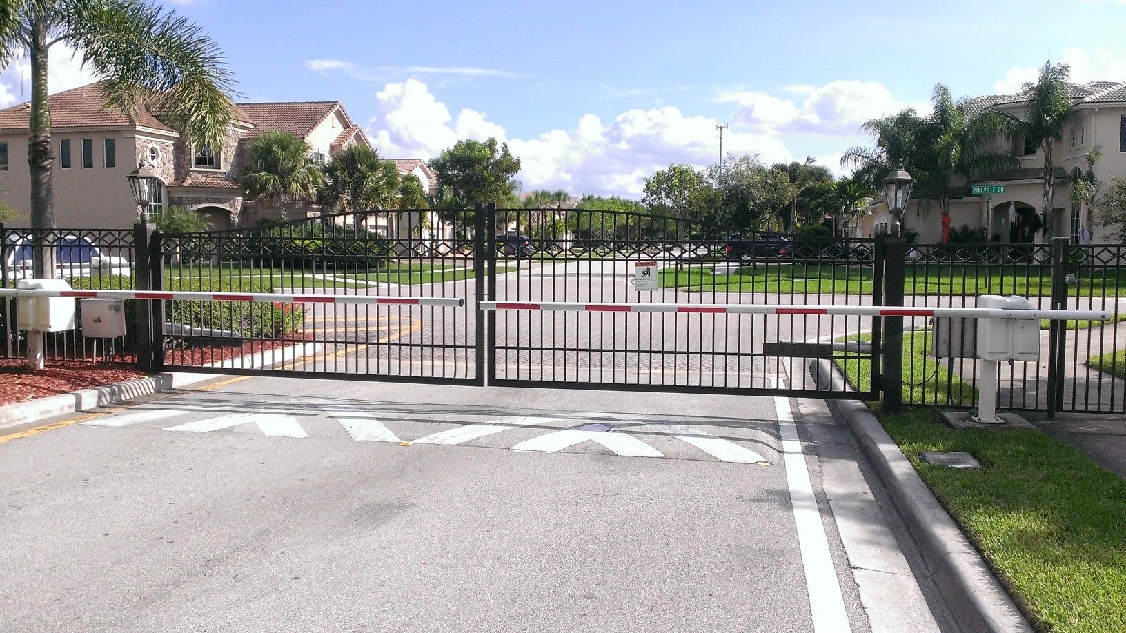 Gated entrance to a residential complex with palm trees, buildings, and a closed black metal gate.