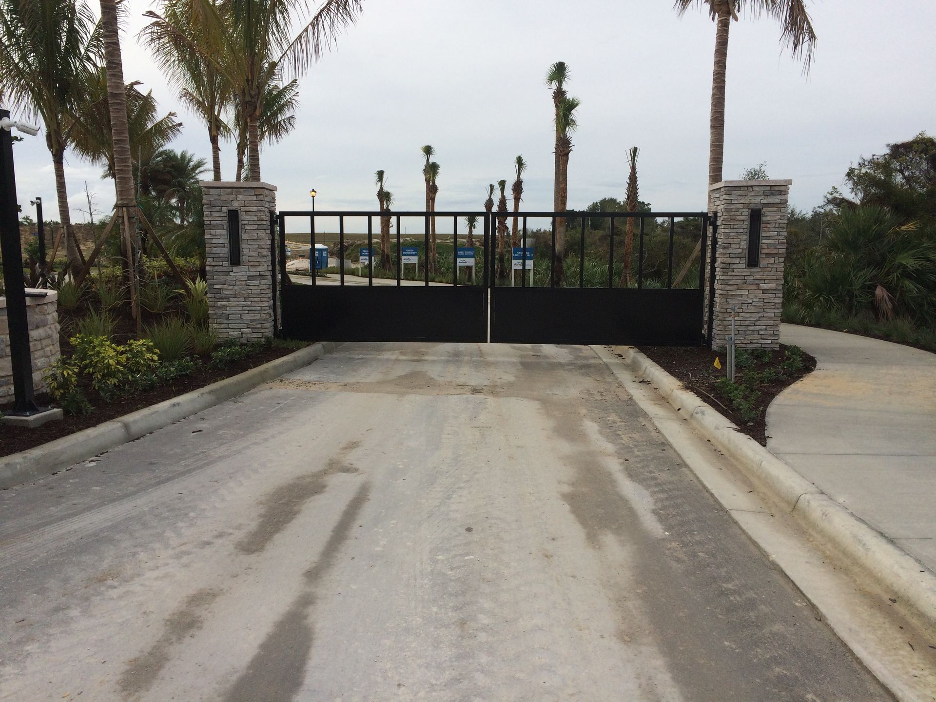 Closed black gate at the end of a driveway, flanked by stone pillars and palm trees