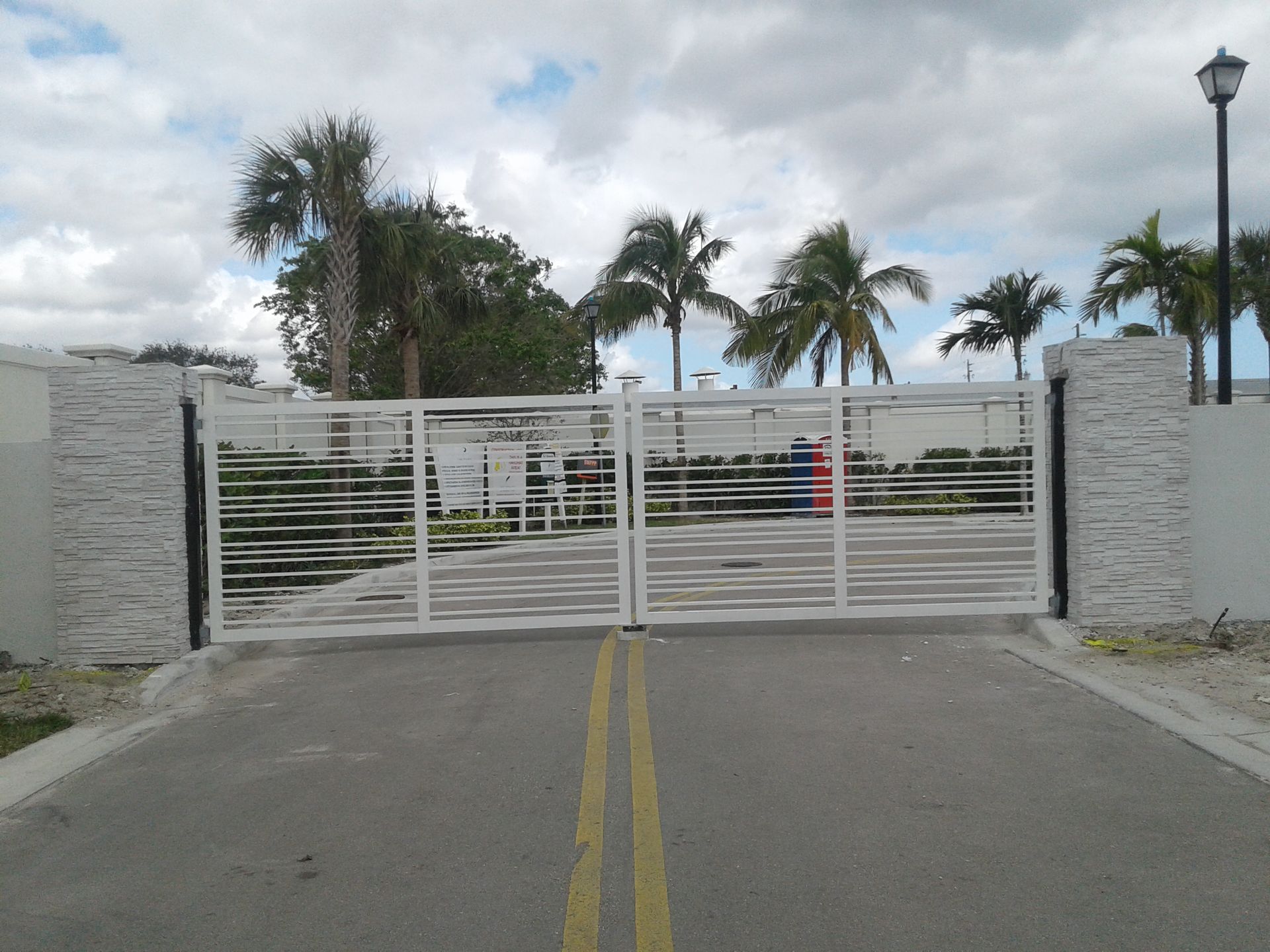 Closed white gate at the end of a road, with palm trees and a lamppost behind it.