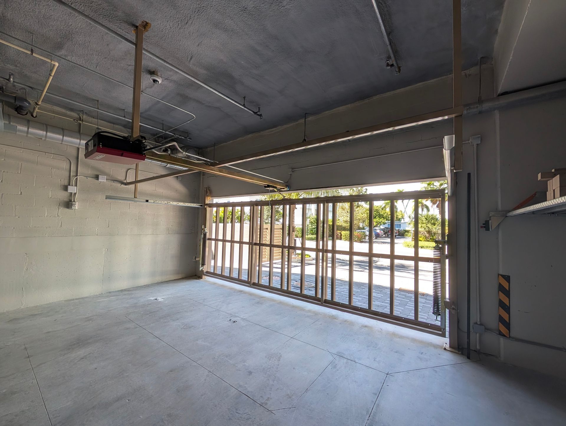Empty garage interior with concrete floor, exposed ceiling, and open metal-framed door to outside