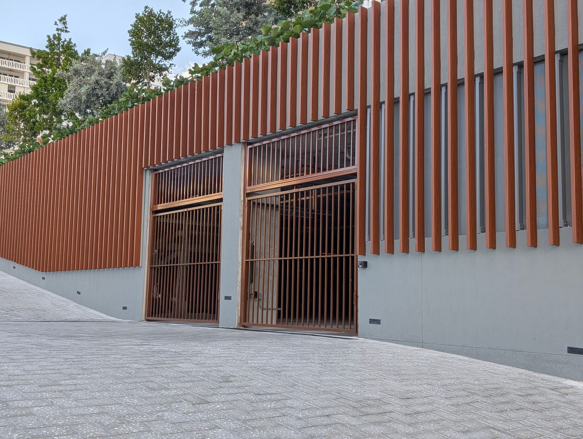 Modern building entrance with rust-colored slatted facade and large metal gates on a sloped paved street