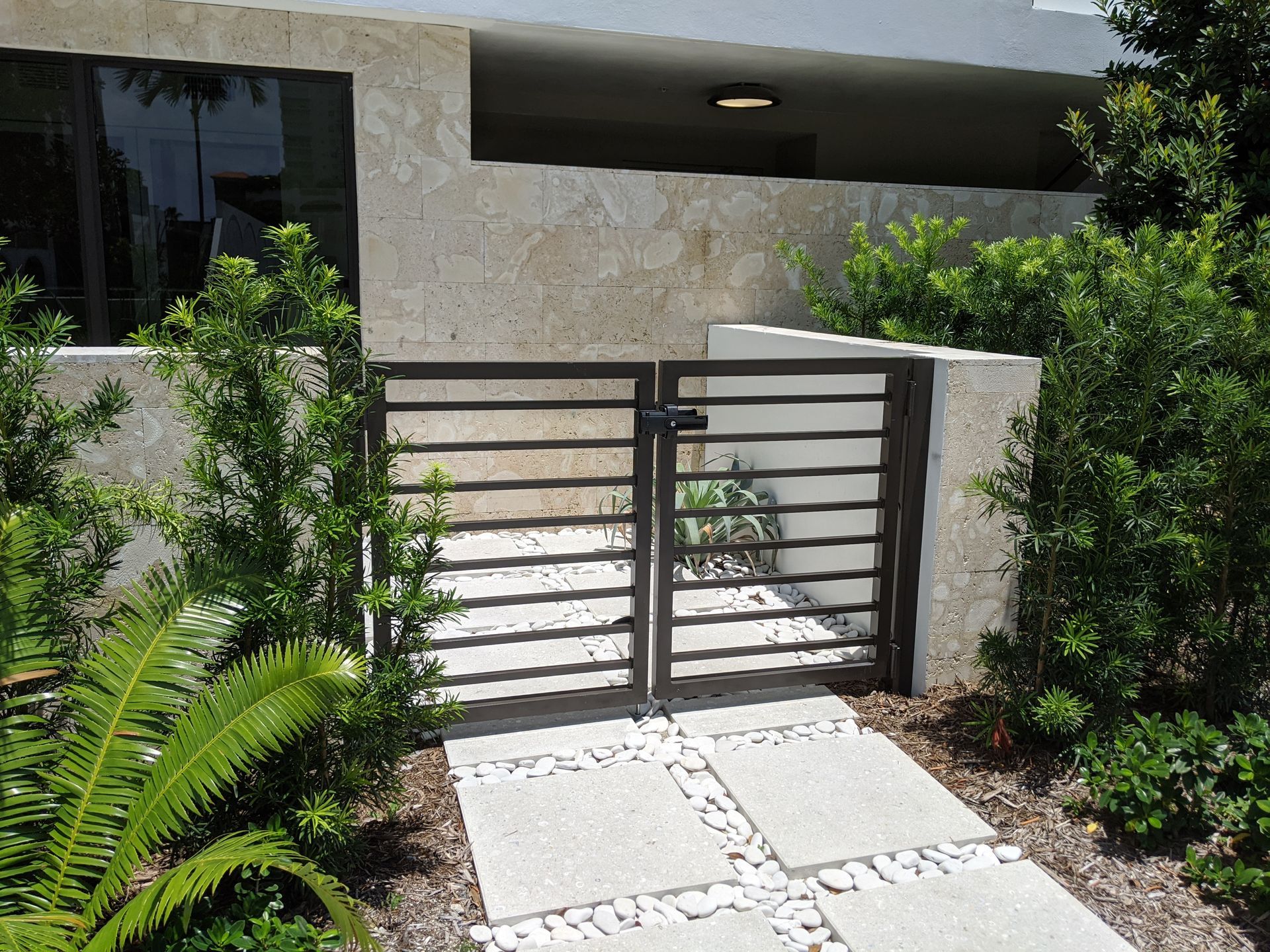 Modern house entry gate with black metal slats, stone wall, and landscaped greenery along a paved walkway