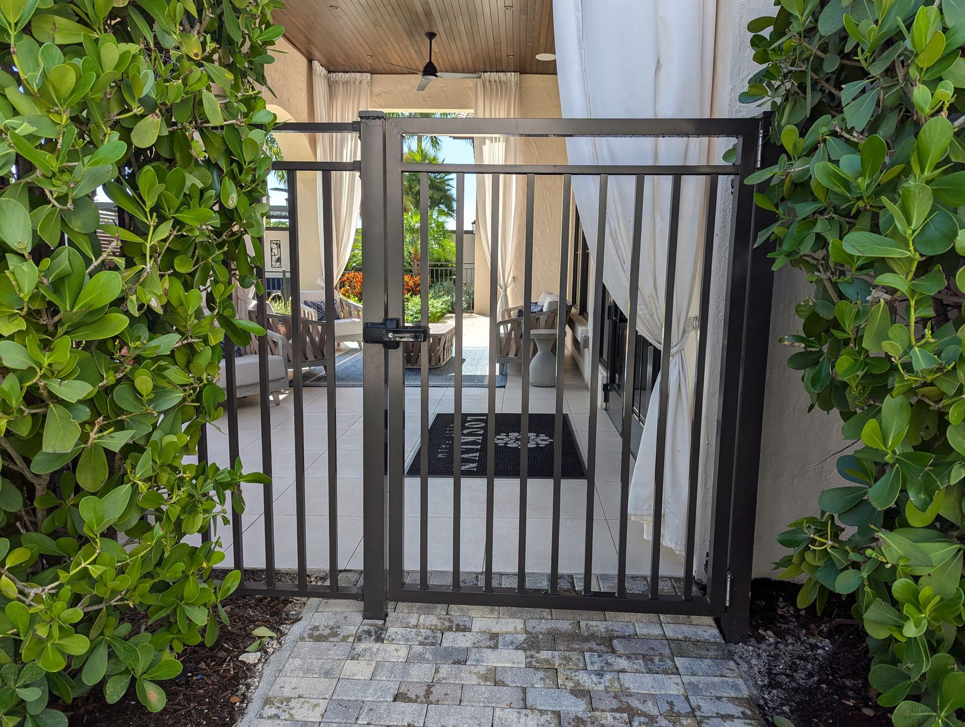 Black metal gate at a stone patio entrance framed by green hedges and draped curtains
