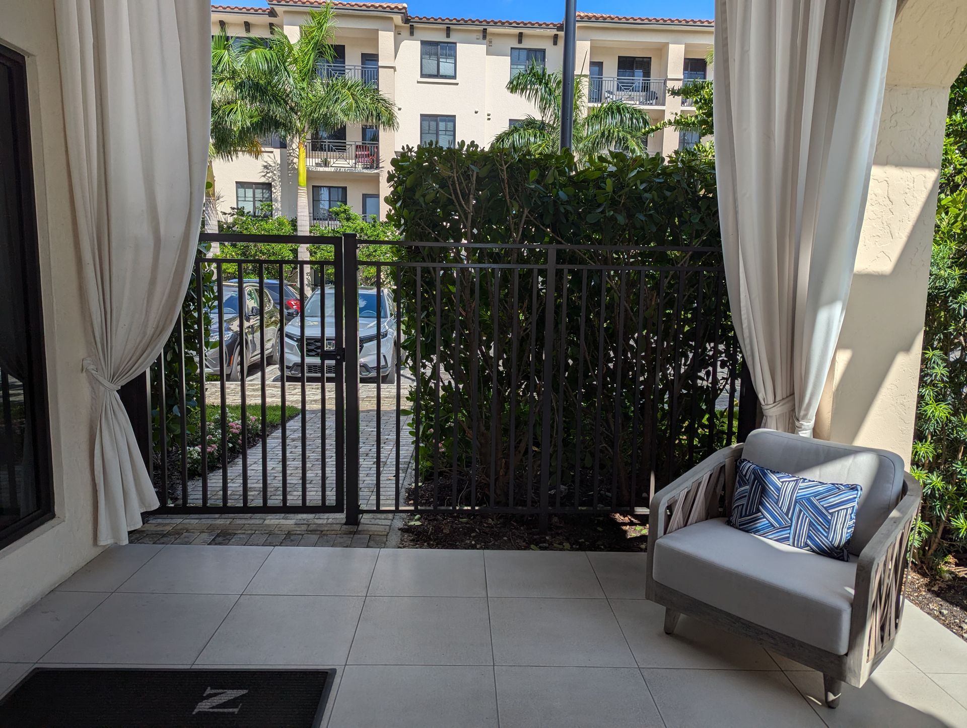 Balcony with black railing, gray tile floor, curtains, and a chair facing apartment buildings and greenery.
