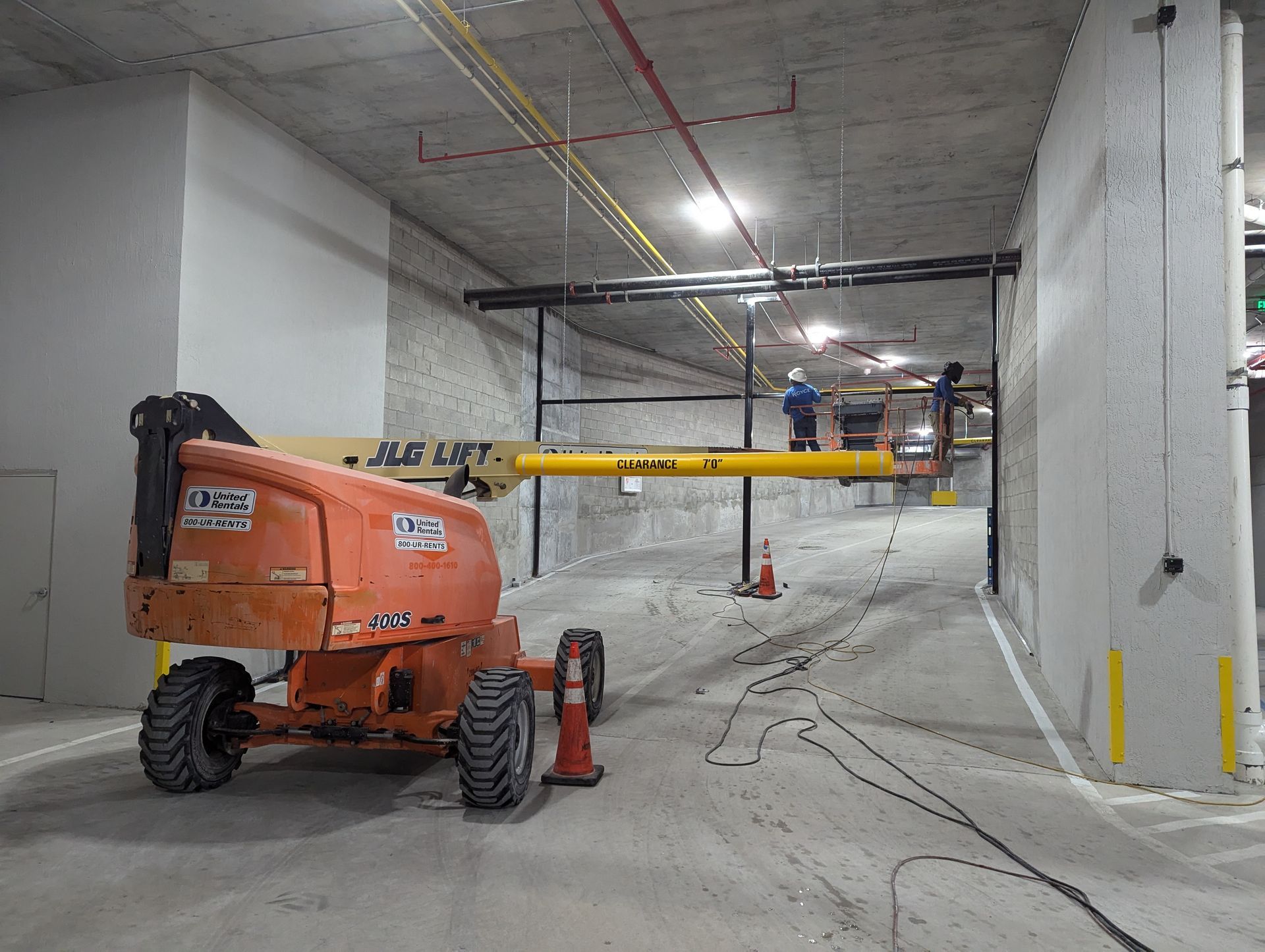 Construction site with orange boom lift in a concrete garage, workers on a yellow lift near the ceiling.
