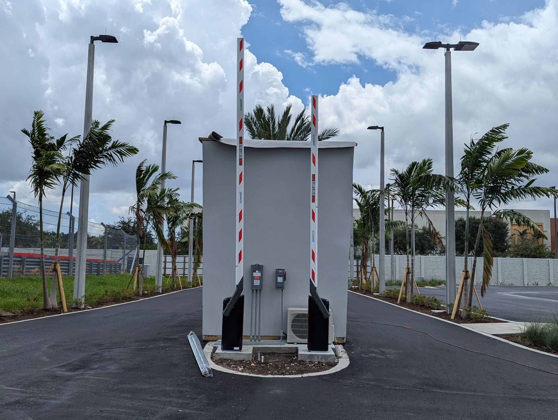 White utility cabinet on a road median, with striped barrier poles, palm trees, and a cloudy sky behind it