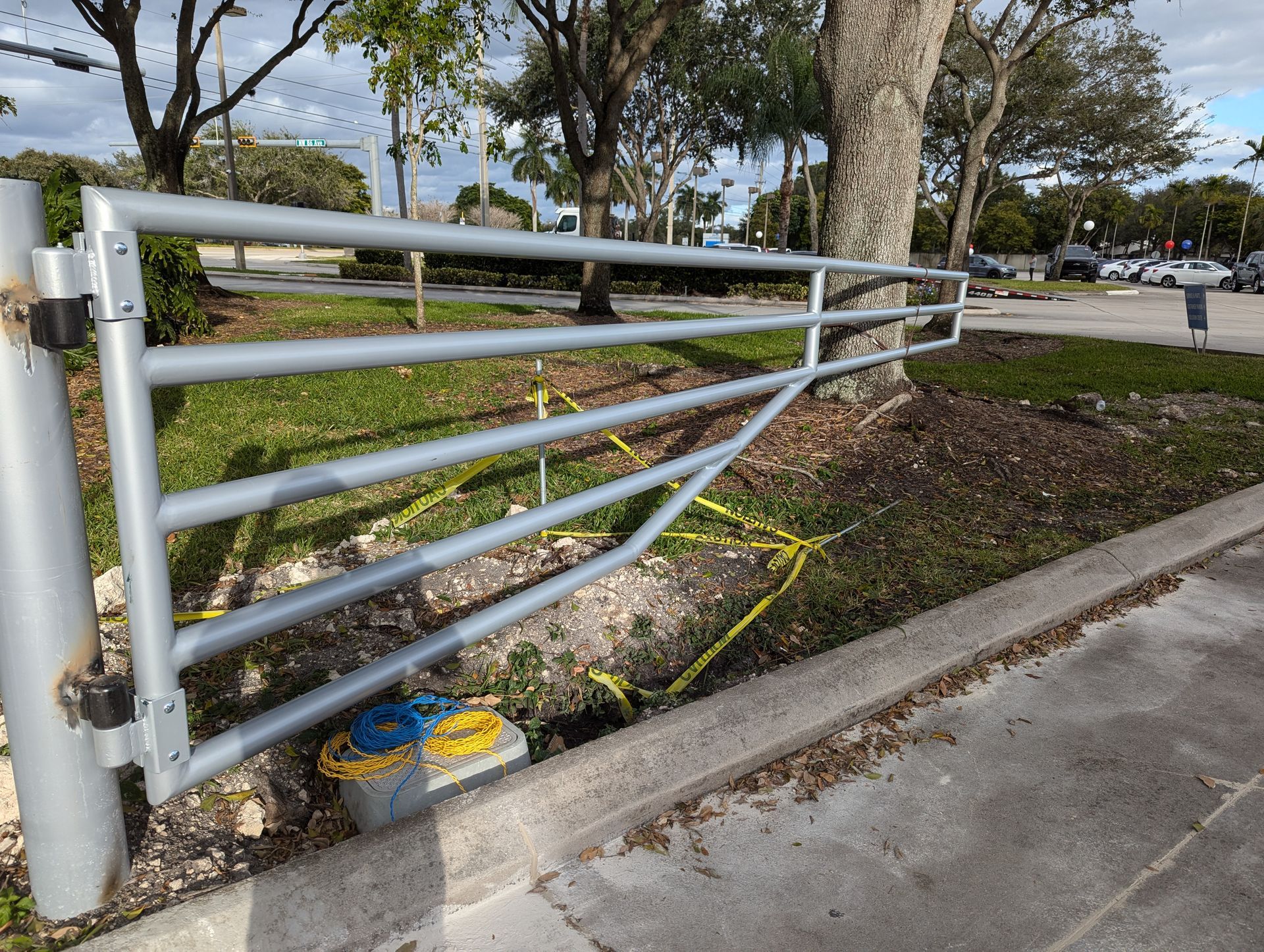 Damaged white metal fence along a sidewalk, bent into a ditch beside a road and trees