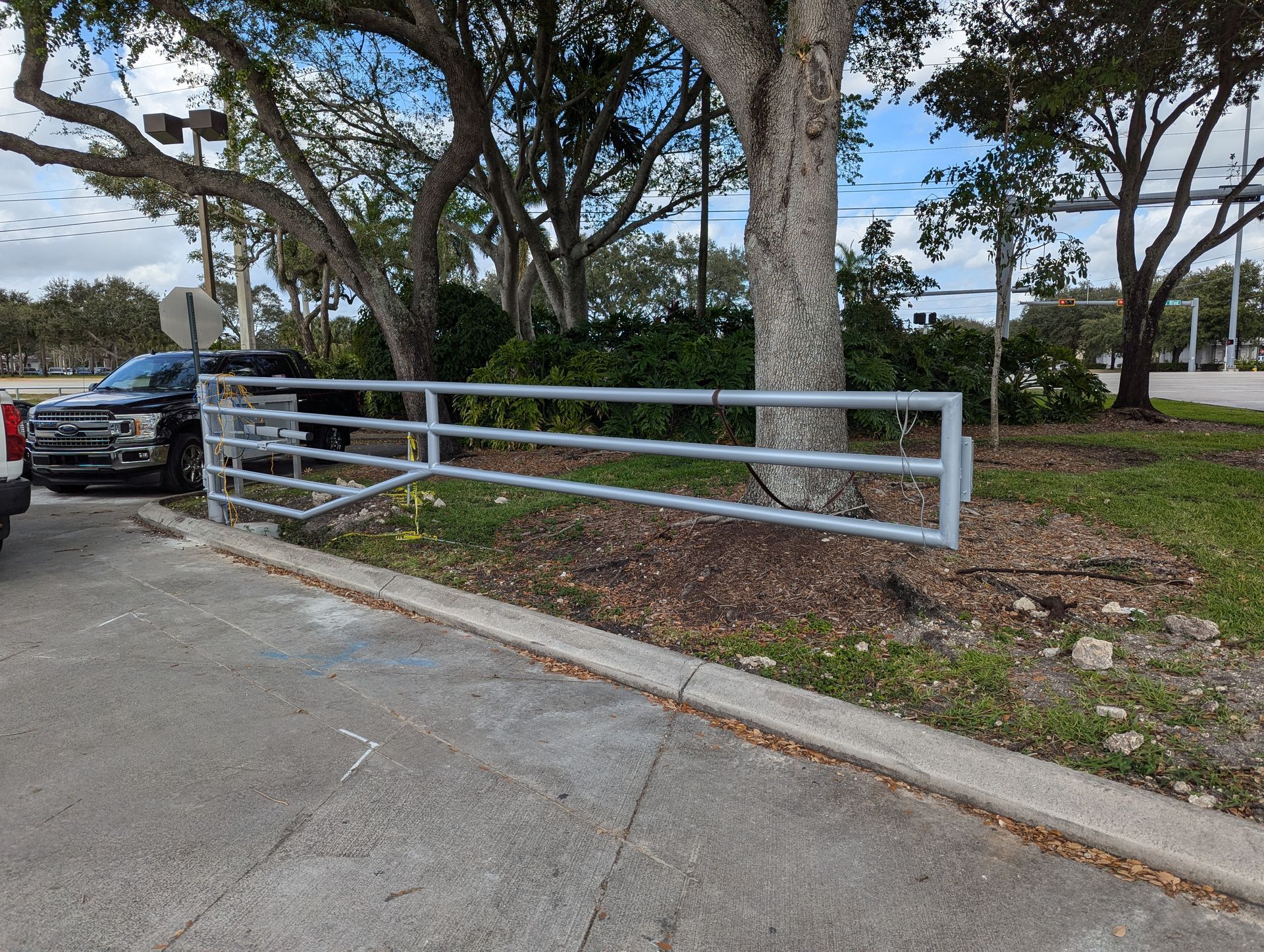 White metal fence beside a parking lot under large trees, with a parked black truck on the left.
