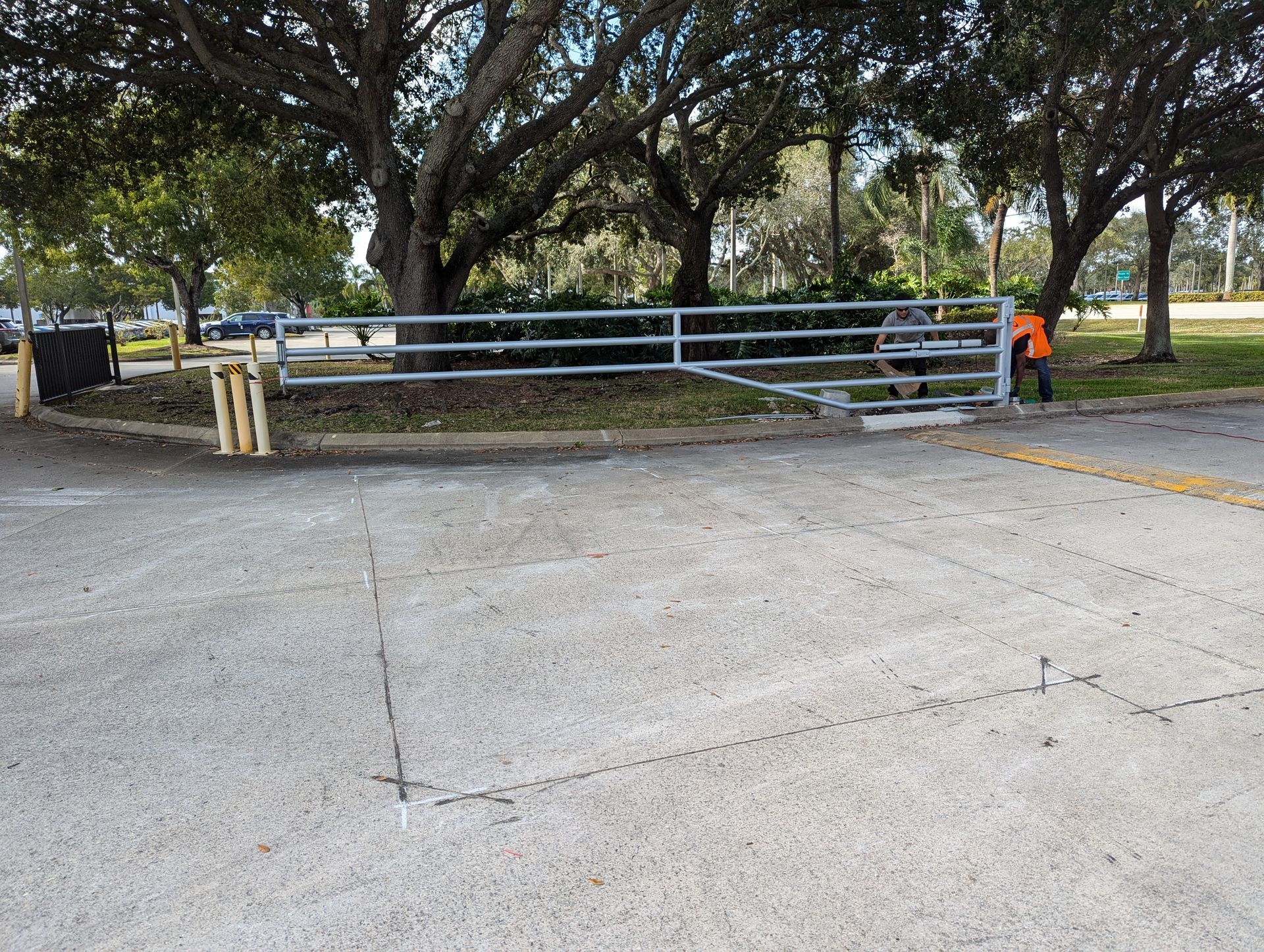 Closed park gate across a concrete driveway, with a worker in orange vest nearby.