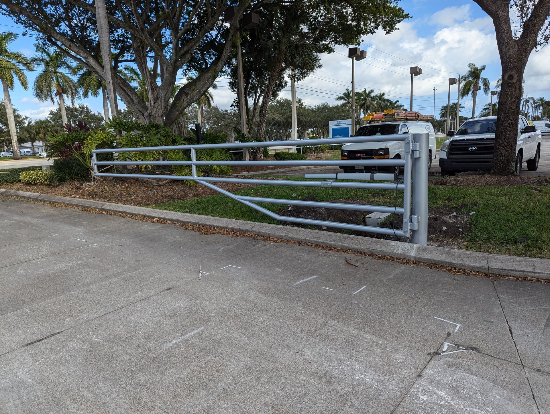 White metal gate across a driveway beside a tree-lined street and parked cars