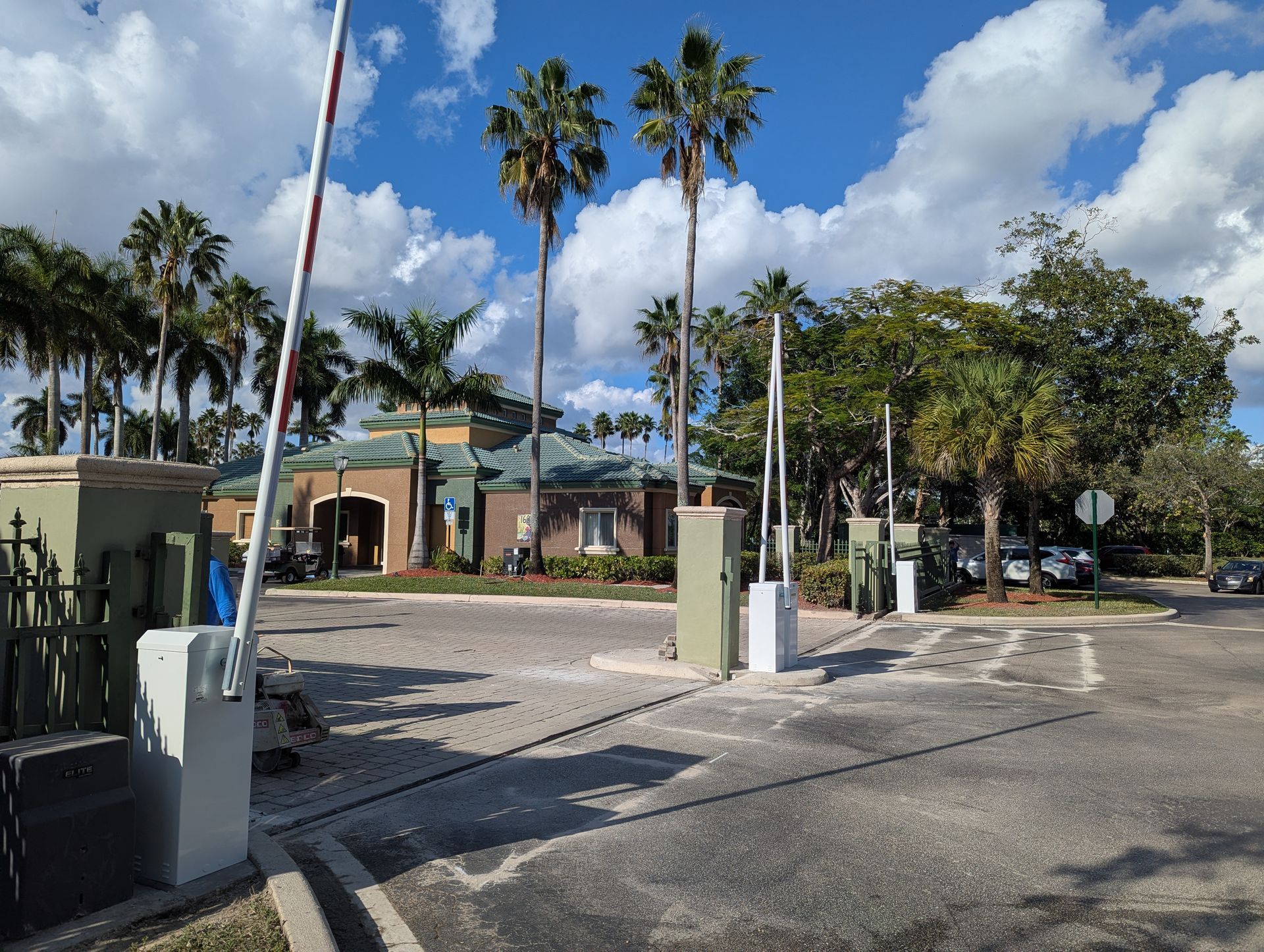 Parking lot entrance with palm trees, a low building, and a raised gate under a partly cloudy sky
