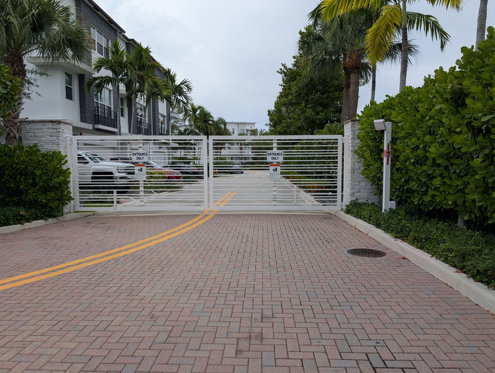 Closed white gate blocking a paved driveway with palm trees and apartment buildings on both sides