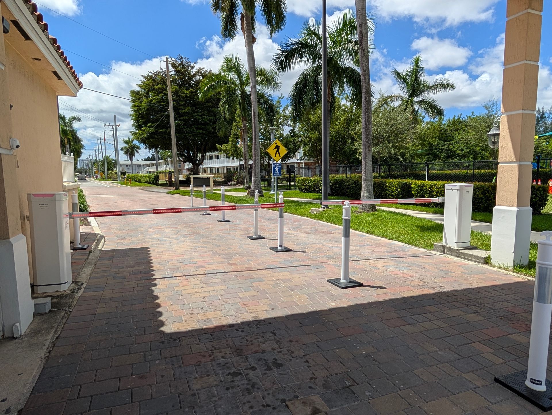 Palm-lined driveway with red and tan pavers, white posts, and a red barrier under a blue sky