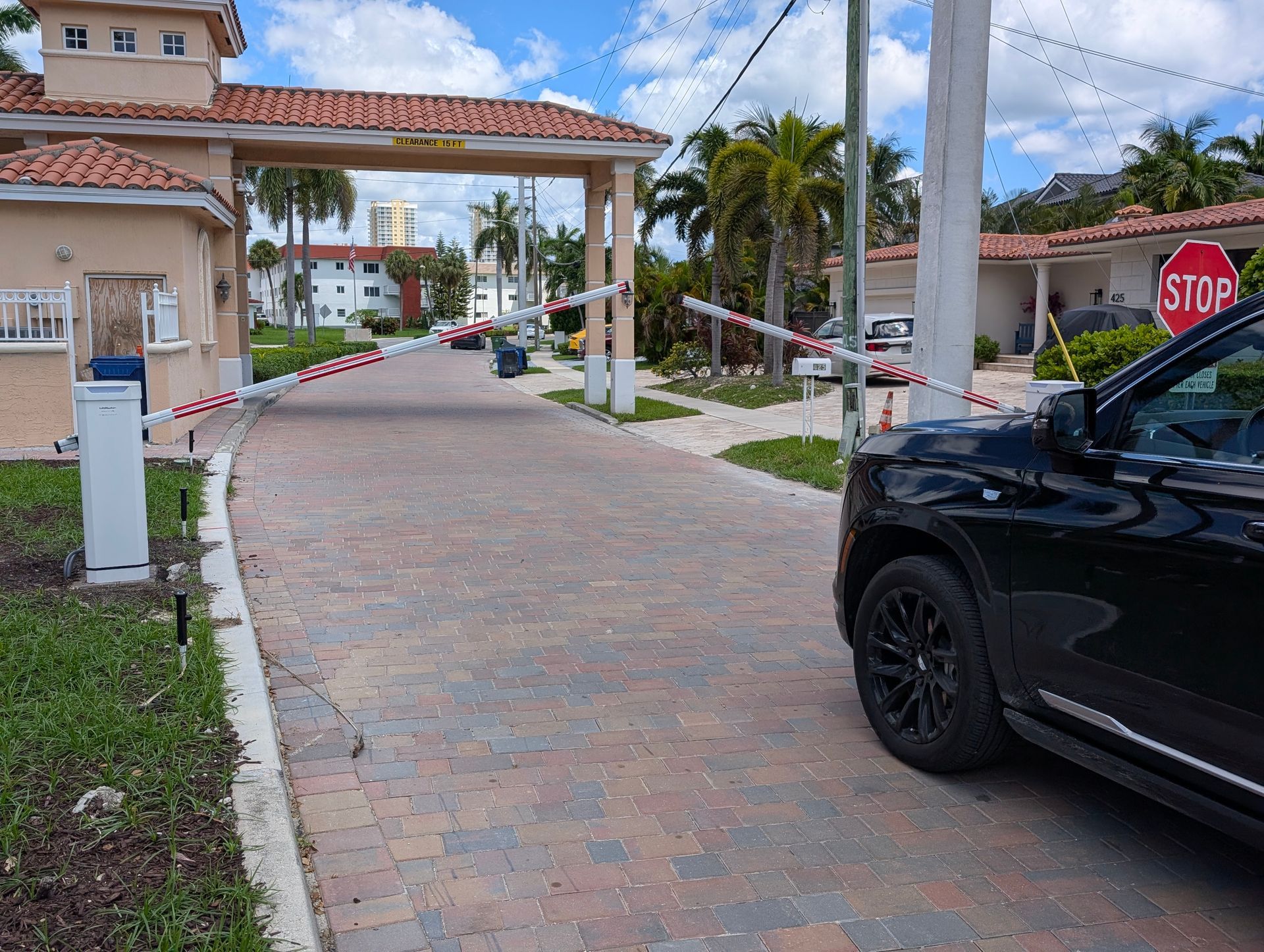 Gated driveway with a black car, stop sign, and tropical buildings under a red-roofed archway