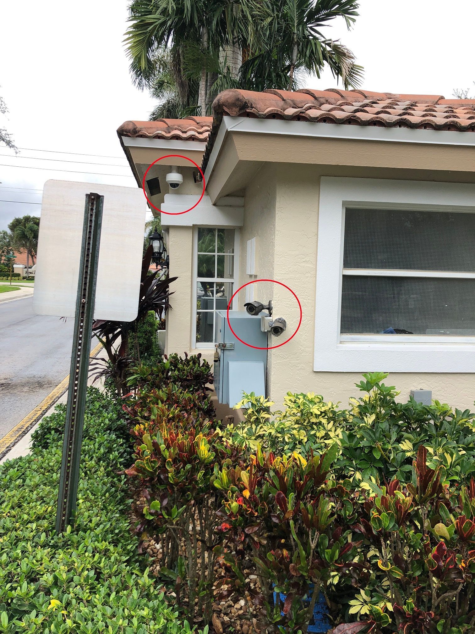 House corner with two security cameras marked by red circles, beside a window and roadside sign.