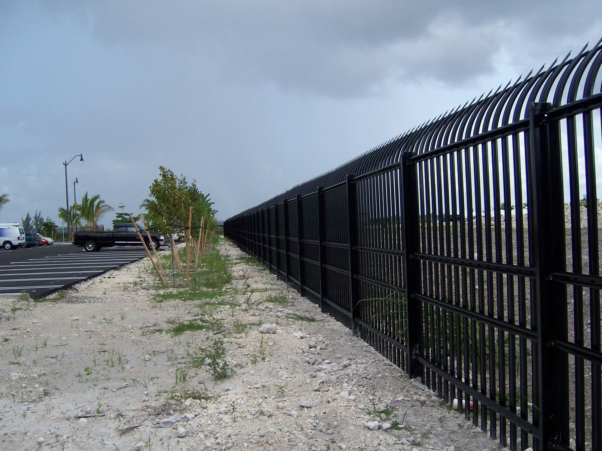Long black metal fence along a gravel path beside a parking lot under a cloudy sky