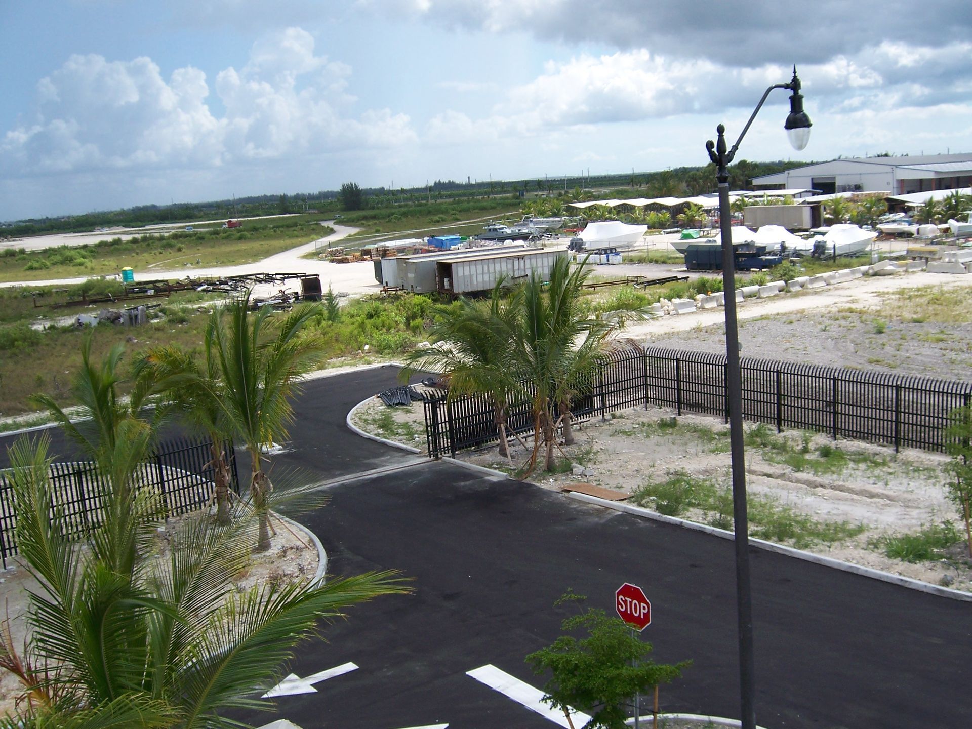 Paved road in a tropical development site with palm trees, lamp posts, and scattered buildings under cloudy skies