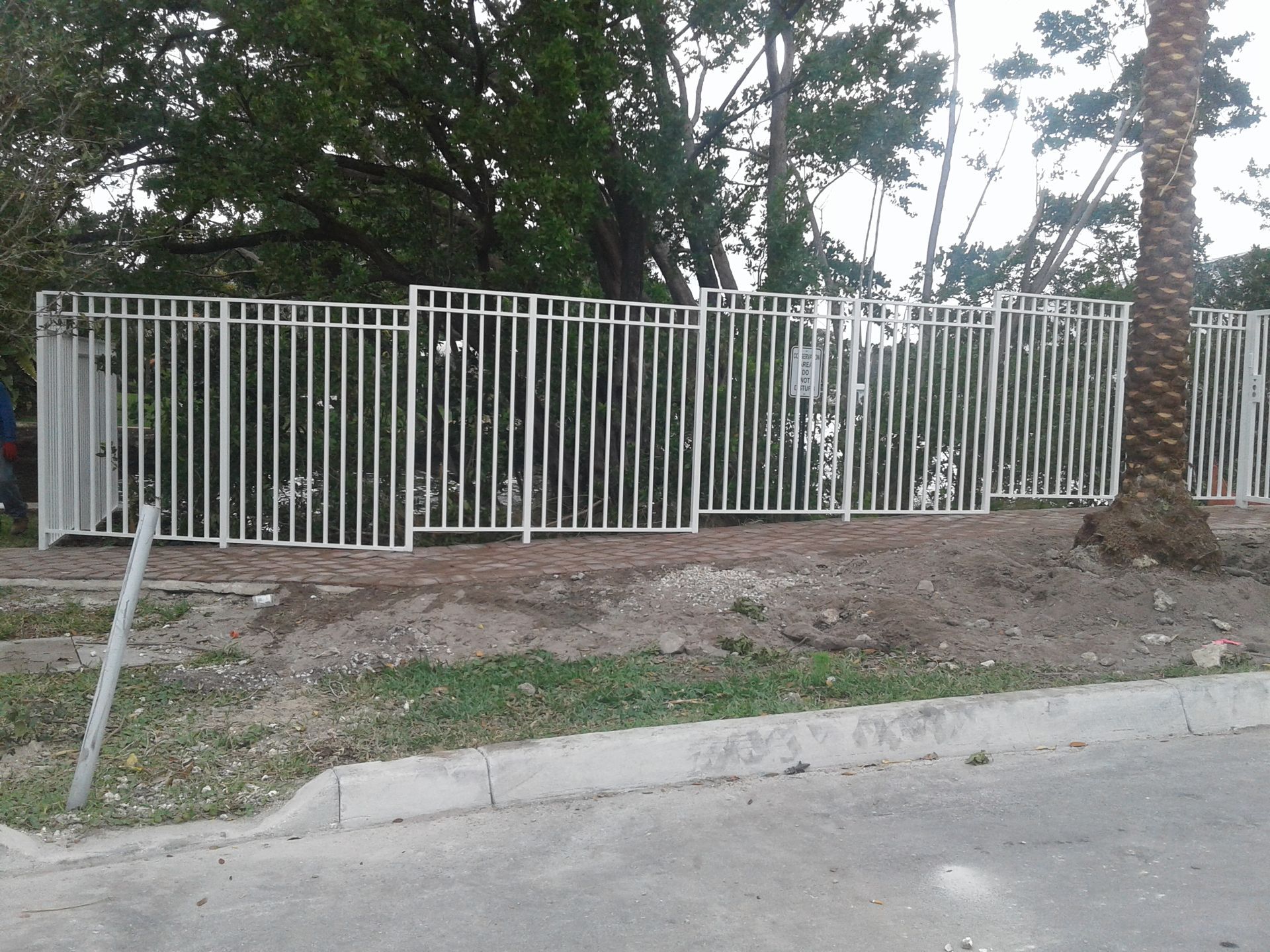 White metal fence along a dirt strip beside a road, with trees behind it