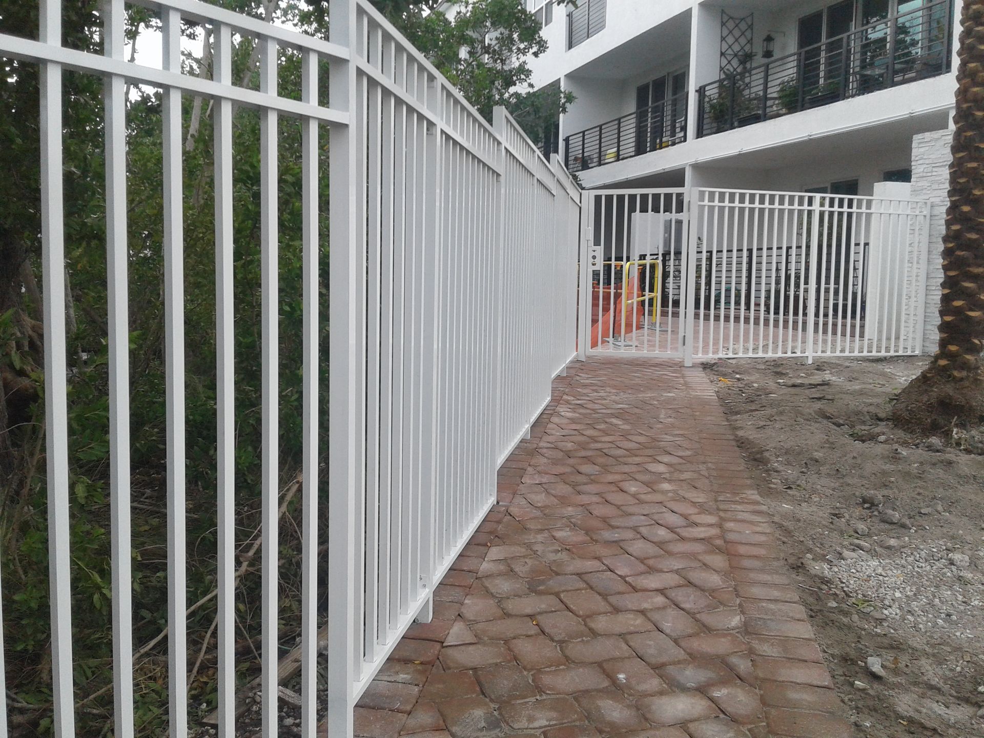 White metal fence along a brick path beside a building and dirt lot
