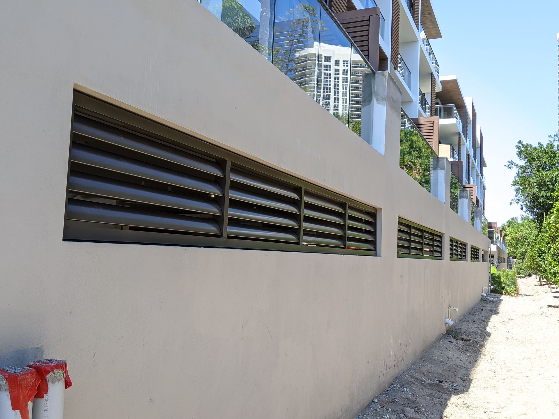 Long beige wall with louvered vents beside a narrow sidewalk on a sunny street
