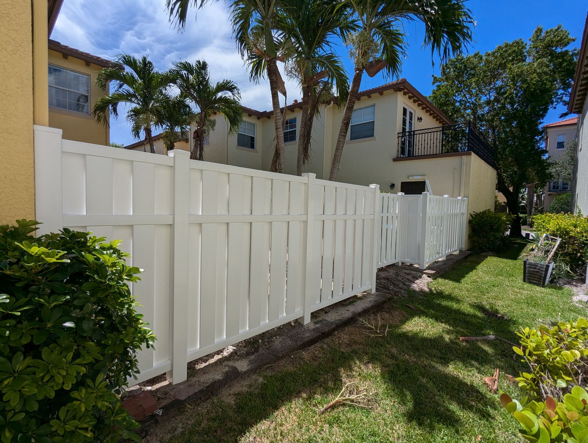 White fence beside a grassy yard with palm trees and beige homes on a sunny day
