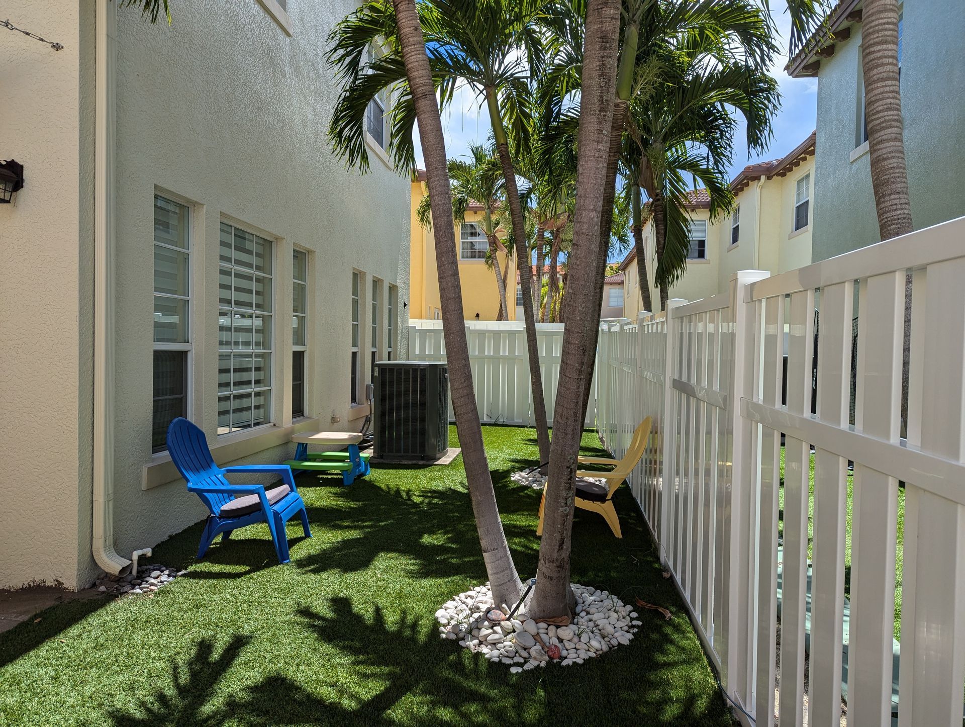 Small courtyard with palm trees, blue chairs, green grass, and a white fence beside a building.
