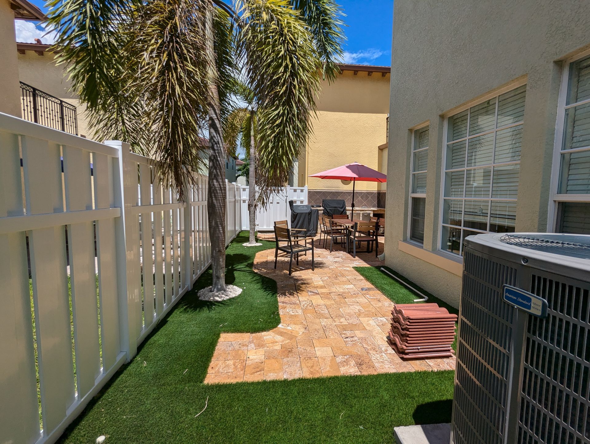 Narrow patio walkway with palm tree, tan pavers, lounge chairs, and a red umbrella between buildings
