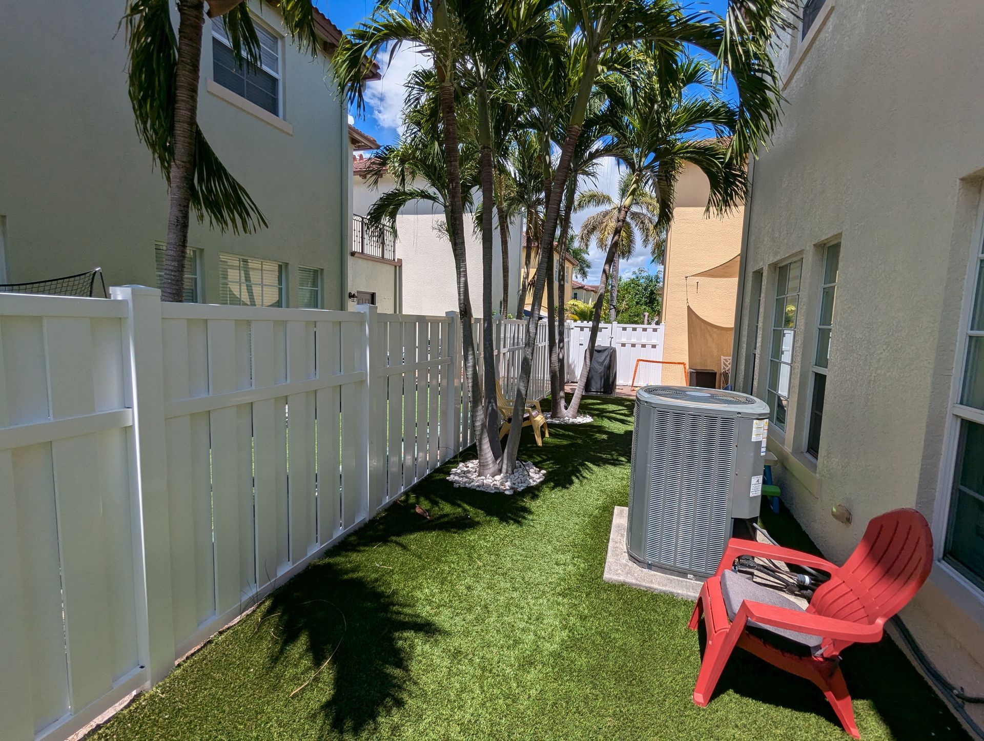 Narrow outdoor walkway with white fence, palm trees, and red chair beside a beige building.
