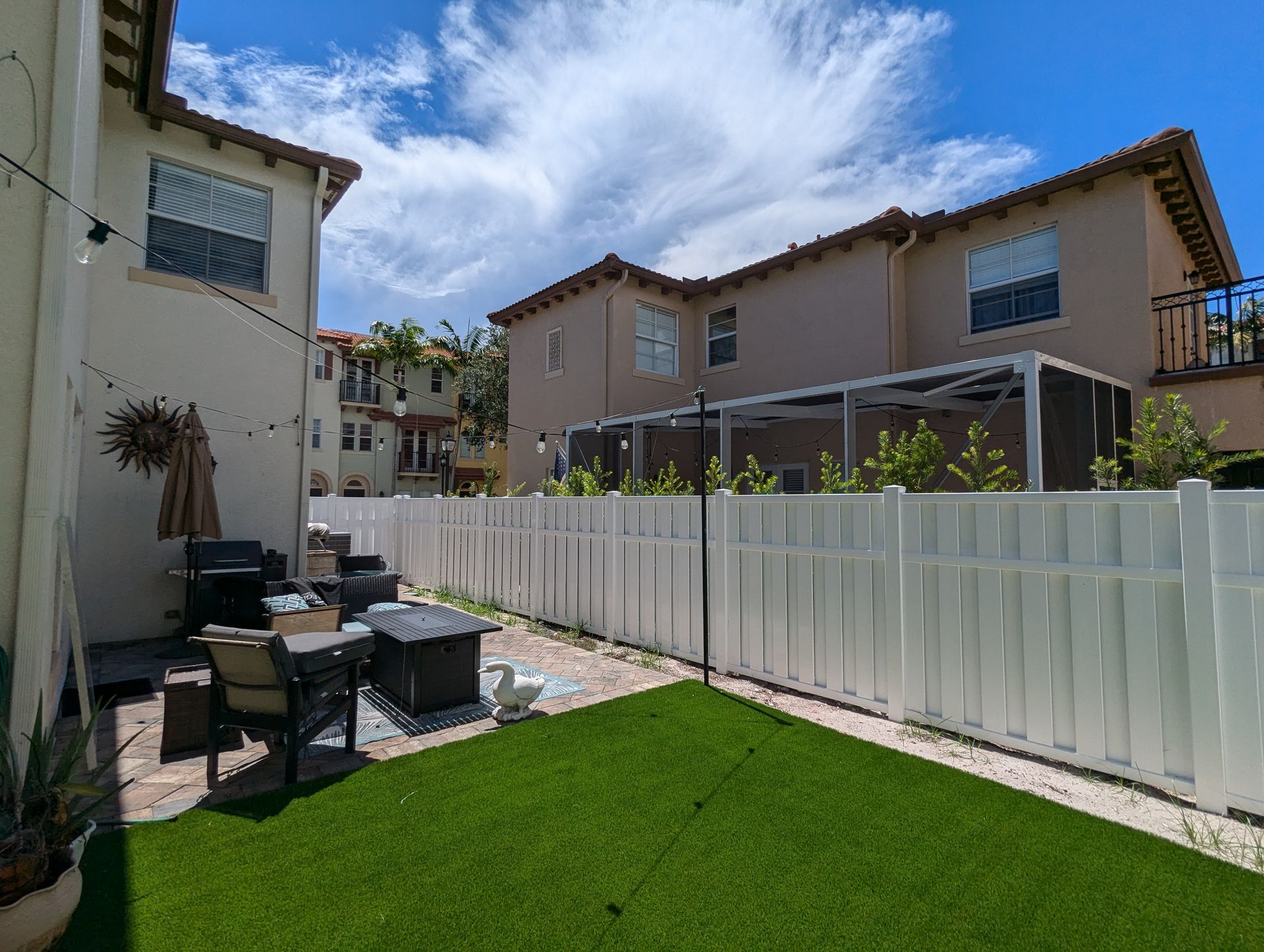 Backyard patio with artificial grass, outdoor chairs, and a white fence beside townhouses.