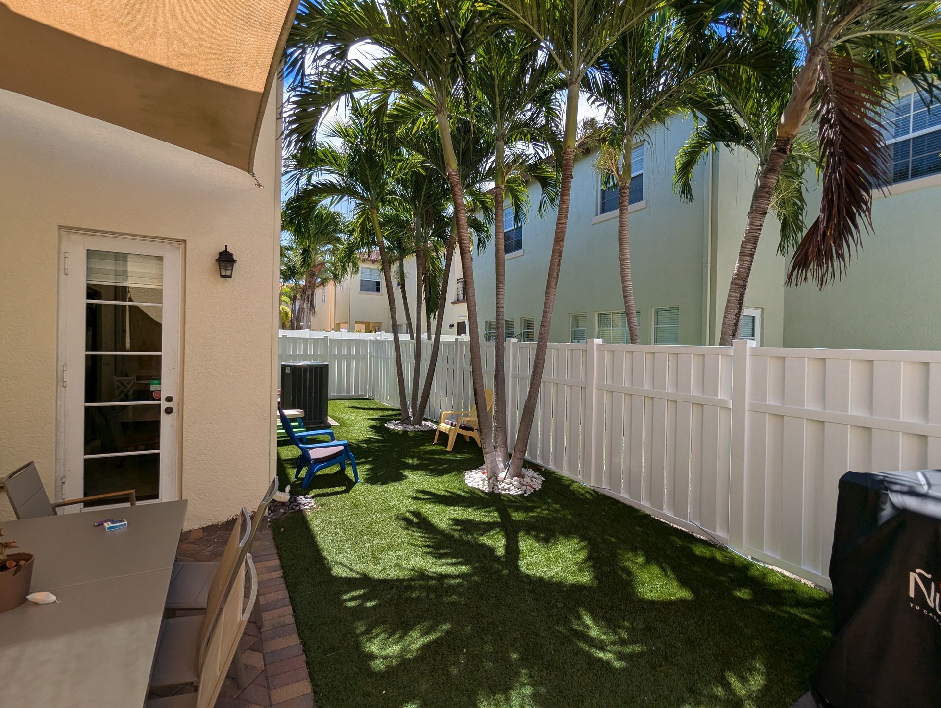 Patio beside a white fence and palm trees, with outdoor chairs and a door to the house.
