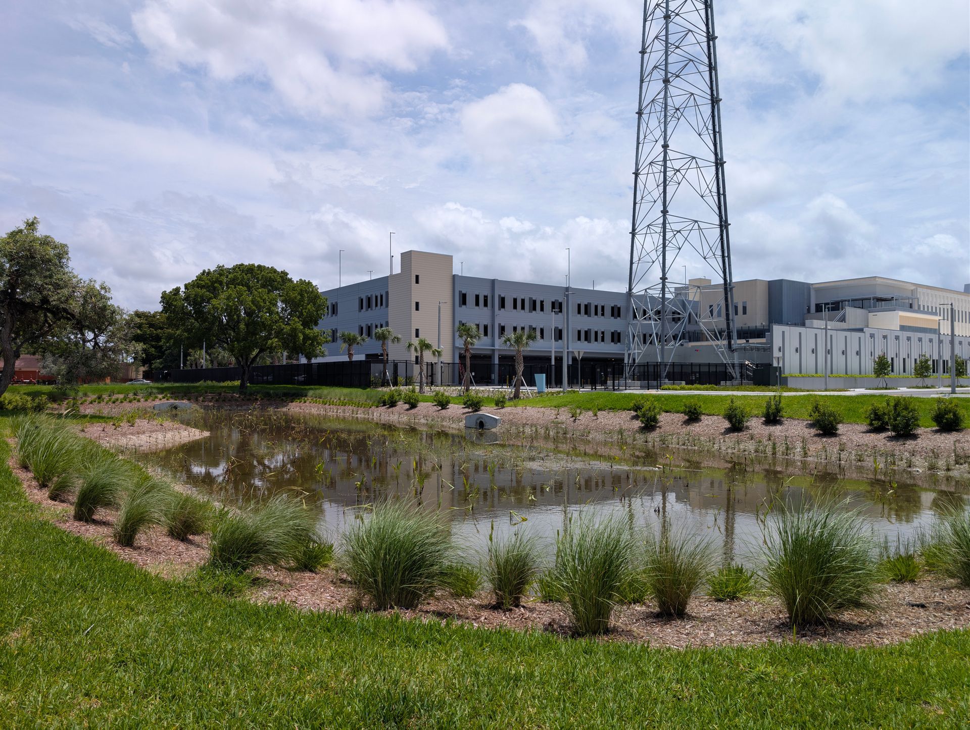 Pond and grassy landscaping in front of a modern industrial building with a tall metal tower under cloudy skies