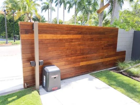 Wooden driveway gate beside a white wall, with a small metal mailbox in front and palm trees beyond.