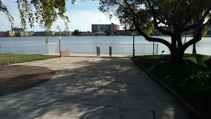 Concrete path leading to a metal fence at a lakeside park, with water, trees, and buildings in the distance