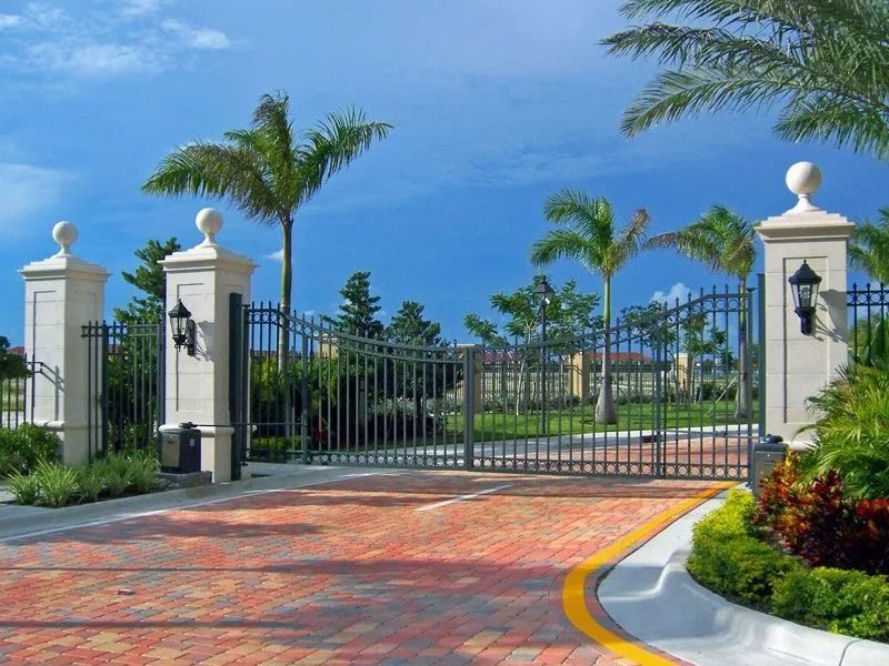 Black wrought-iron gate with white pillars, palm trees, and a brick driveway under a blue sky.