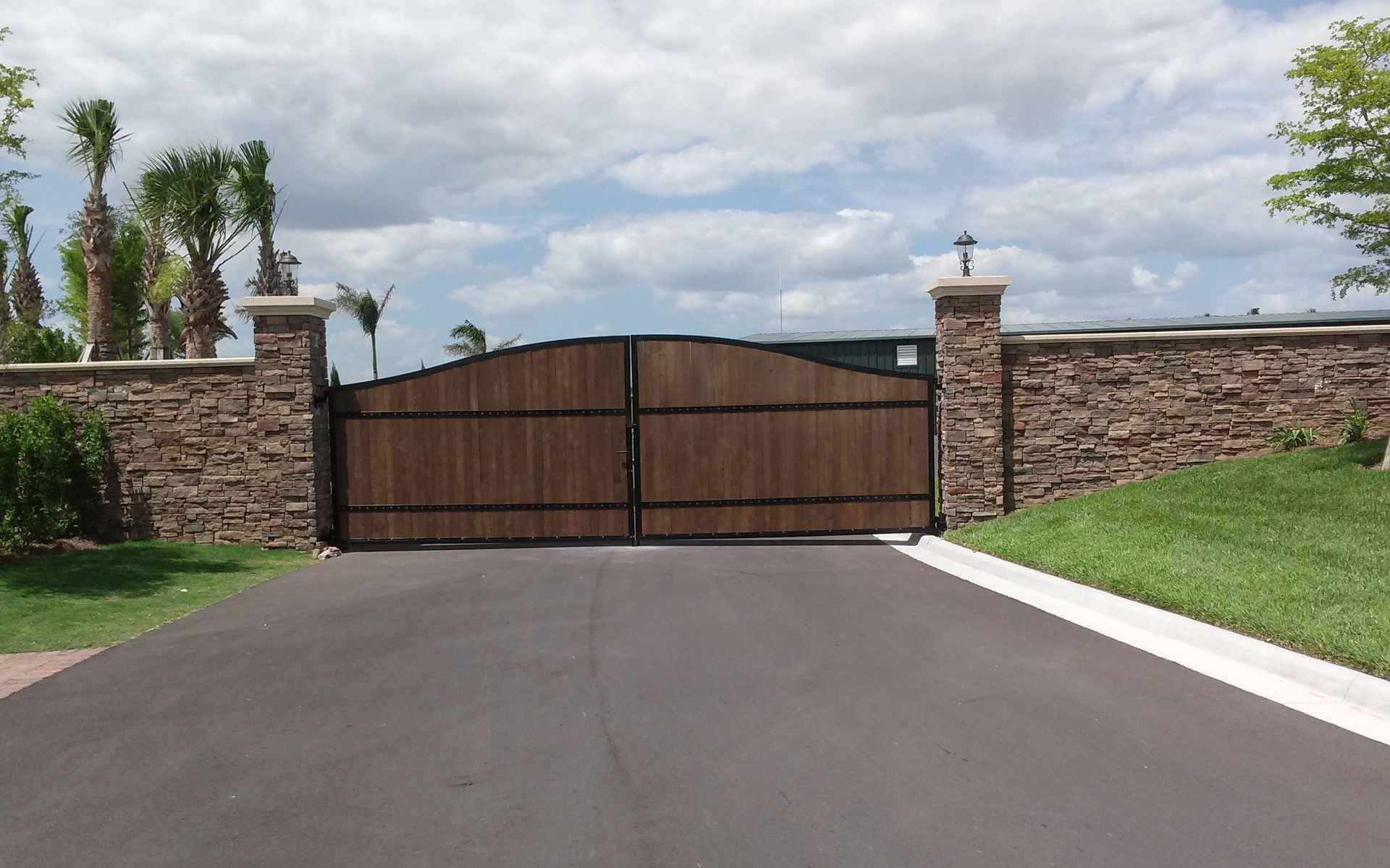 Wooden driveway gate set in a stone wall at the end of a paved road under a cloudy sky