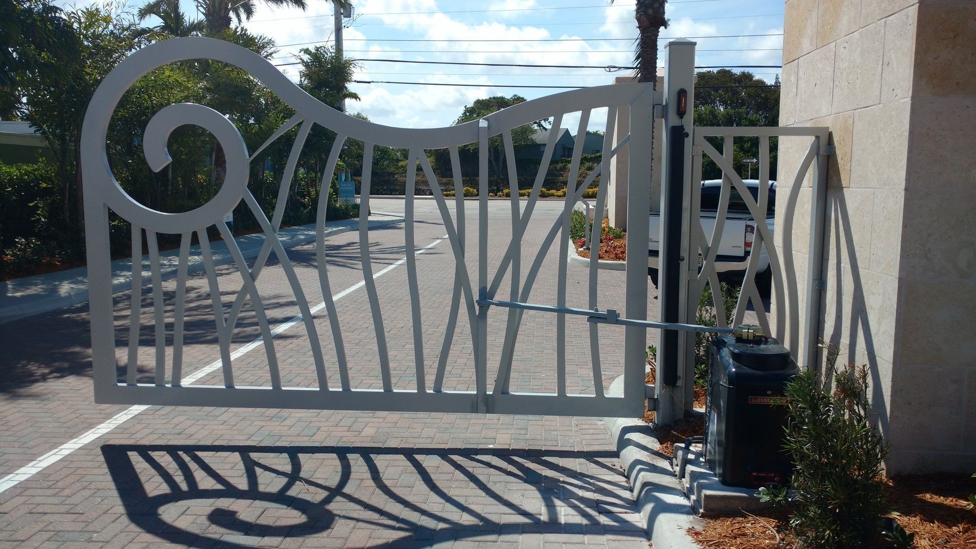 Gray metal driveway gate with spiral design, closed beside a beige wall on a sunny street